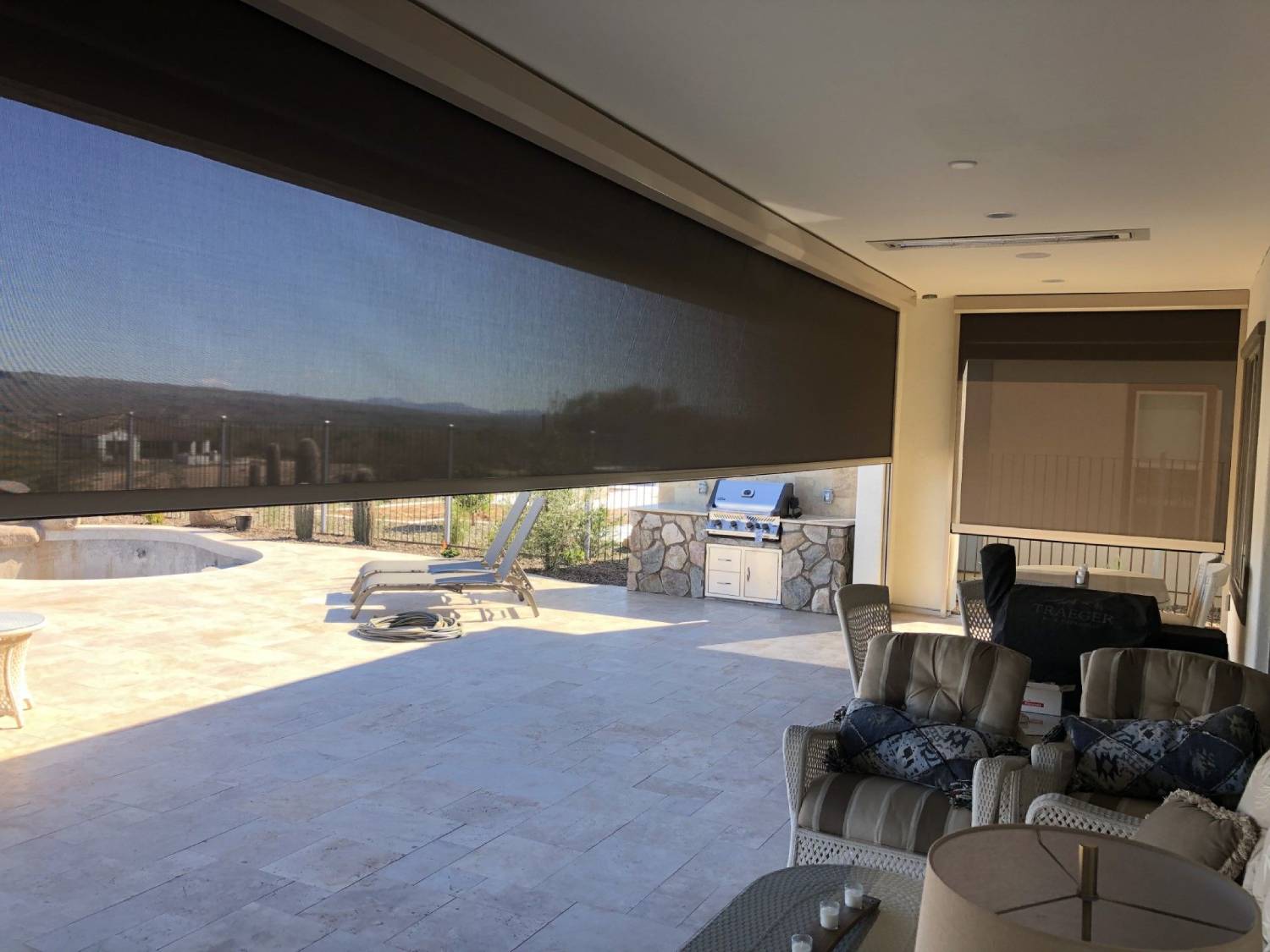 Black solar screen halfway down over a back patio opening. There is patio furniture n the right close to the house and to the left by the outdoor kicthen and pool.