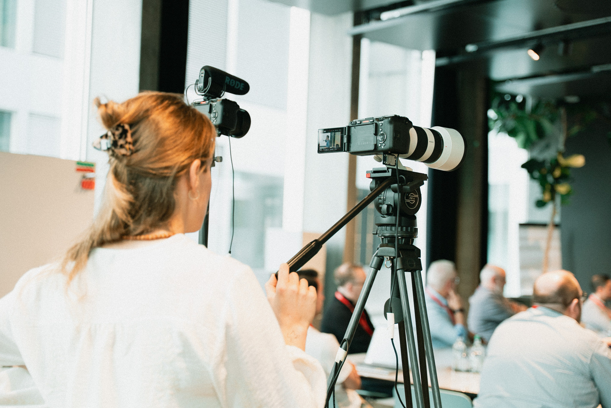 Woman operating a camera on a tripod with a microphone, filming an indoor meeting or conference.