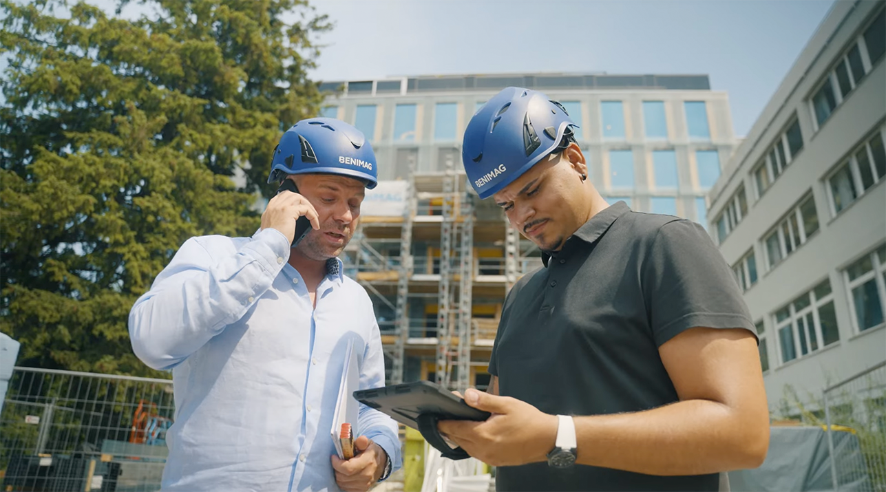 Two men wearing blue BENIMAG helmets, one talking on a phone and the other looking at a tablet, at a construction site.