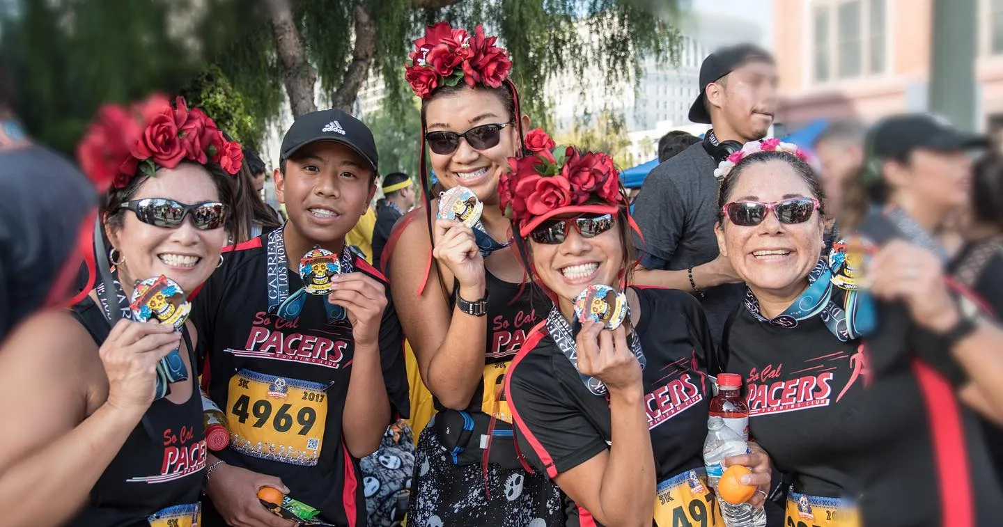 Group of five smiling runners wearing So Cal Pacers shirts and floral headbands, holding medals after a race.