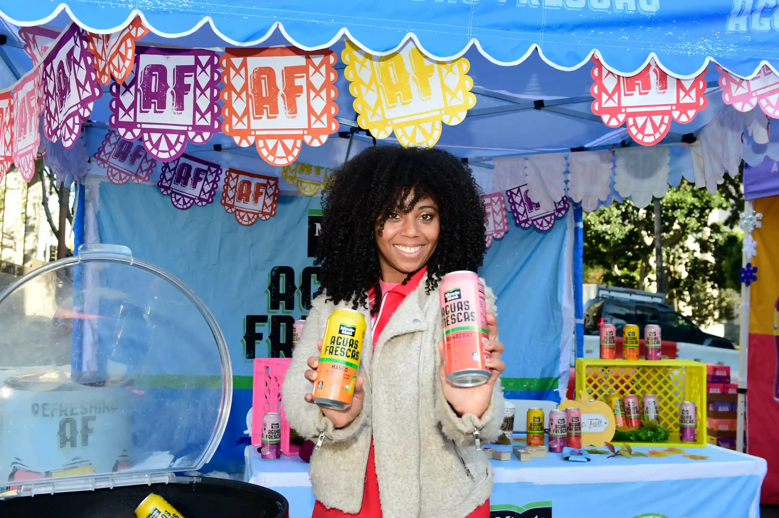 Smiling woman holding two cans of Monte Verde Aguas Frescas beverages at an outdoor booth decorated with colorful banners.