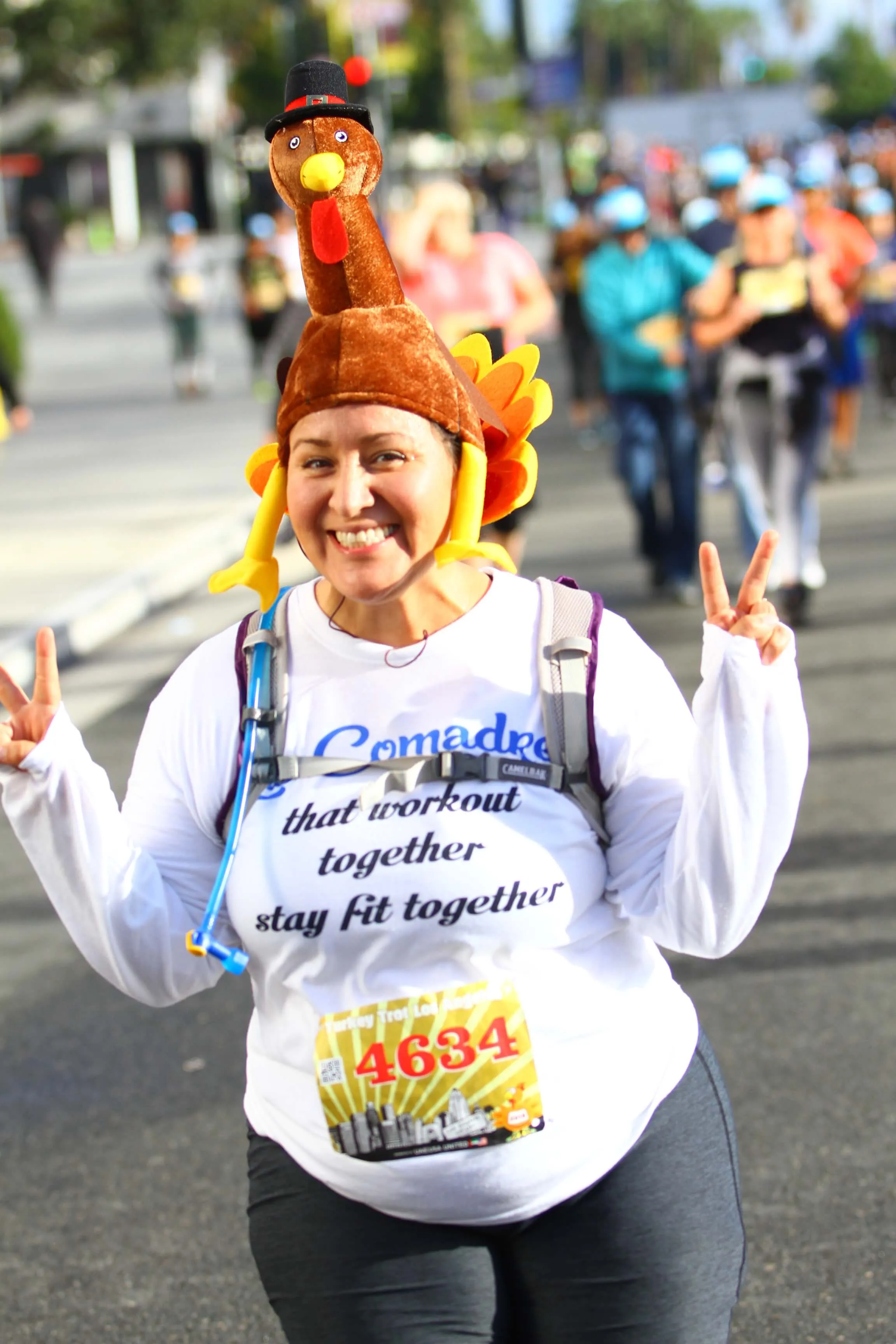 Smiling woman wearing a turkey hat and race bib 4634, making peace signs while participating in a race.