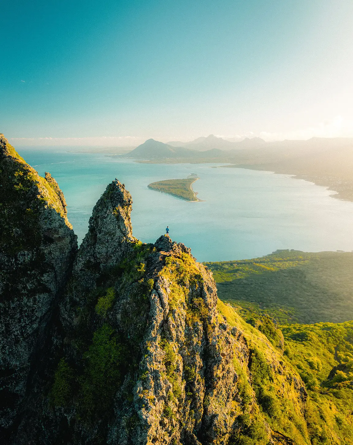 Person standing atop a rocky mountain peak overlooking ocean and island in Mauritius—symbolizing achievement, nature immersion, and elevated travel experiences with Island Lilly