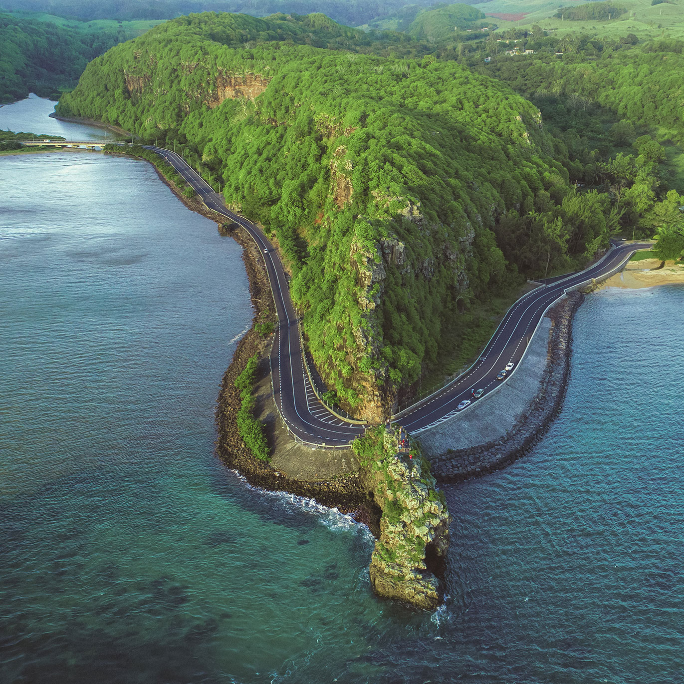 Aerial view of Macondé coastal road winding around a lush cliffside in Mauritius, with ocean and bay below—Island Lilly’s scenic route to discovery and connection
