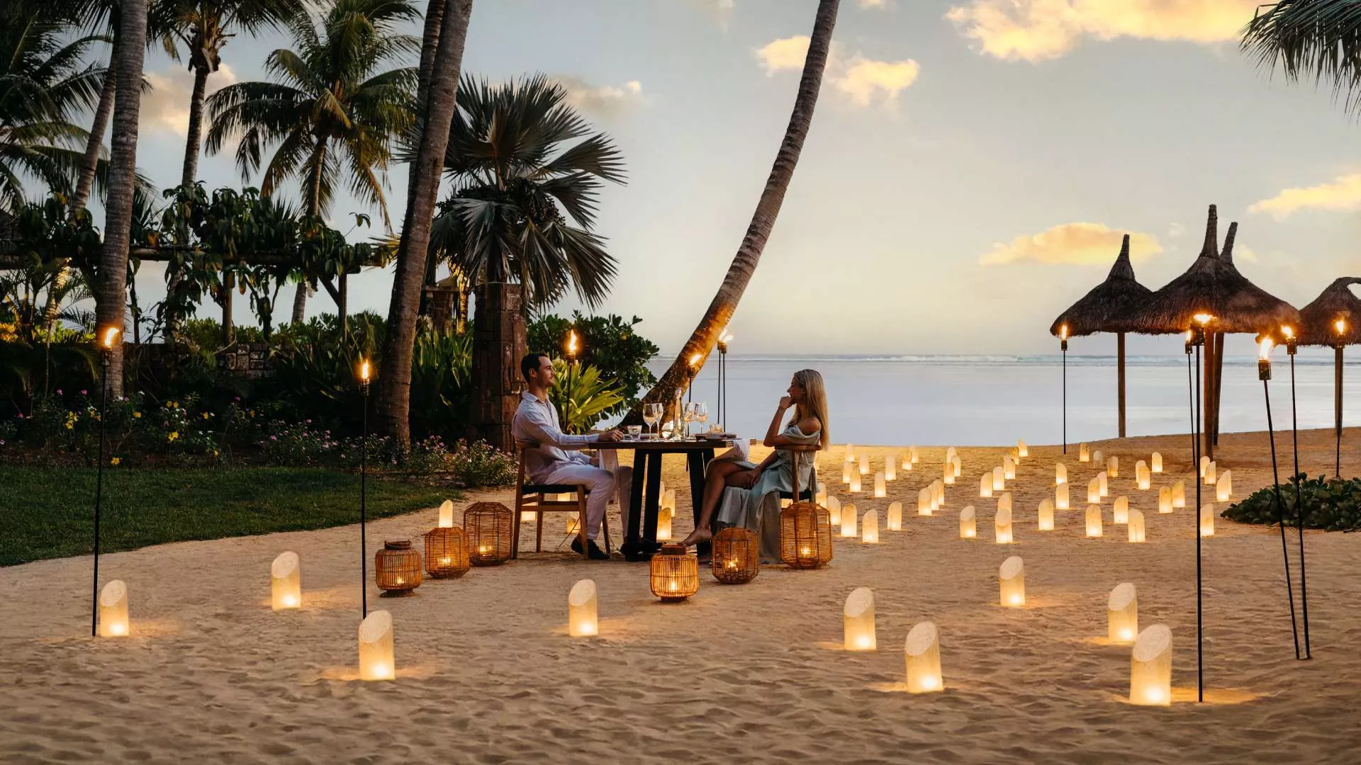 A couple at a candlelit table on the beach at dusk in Mauritius, lanterns around them.