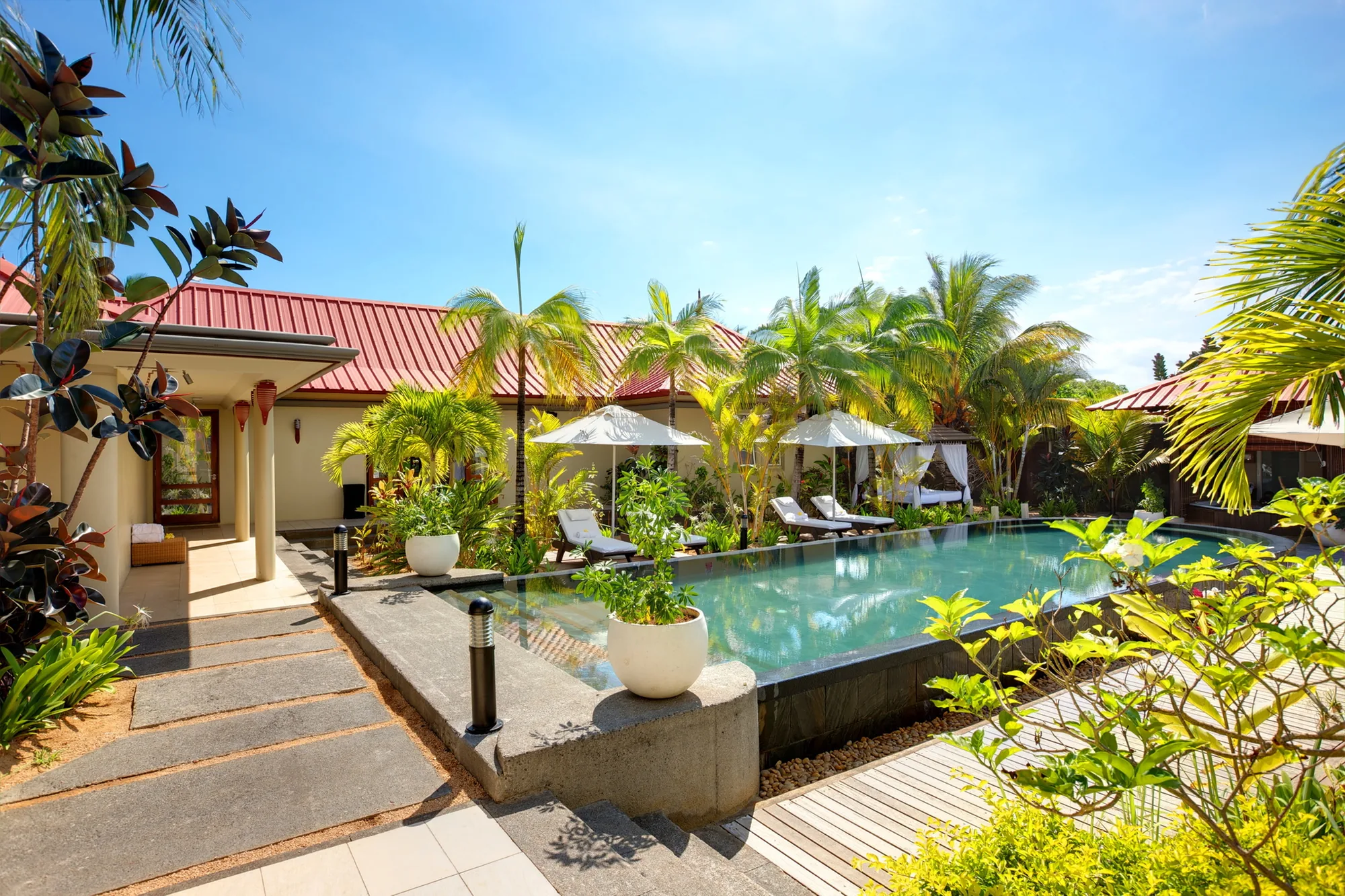 A narrow pool beside red-roofed buildings, with stepping stones and tropical planting