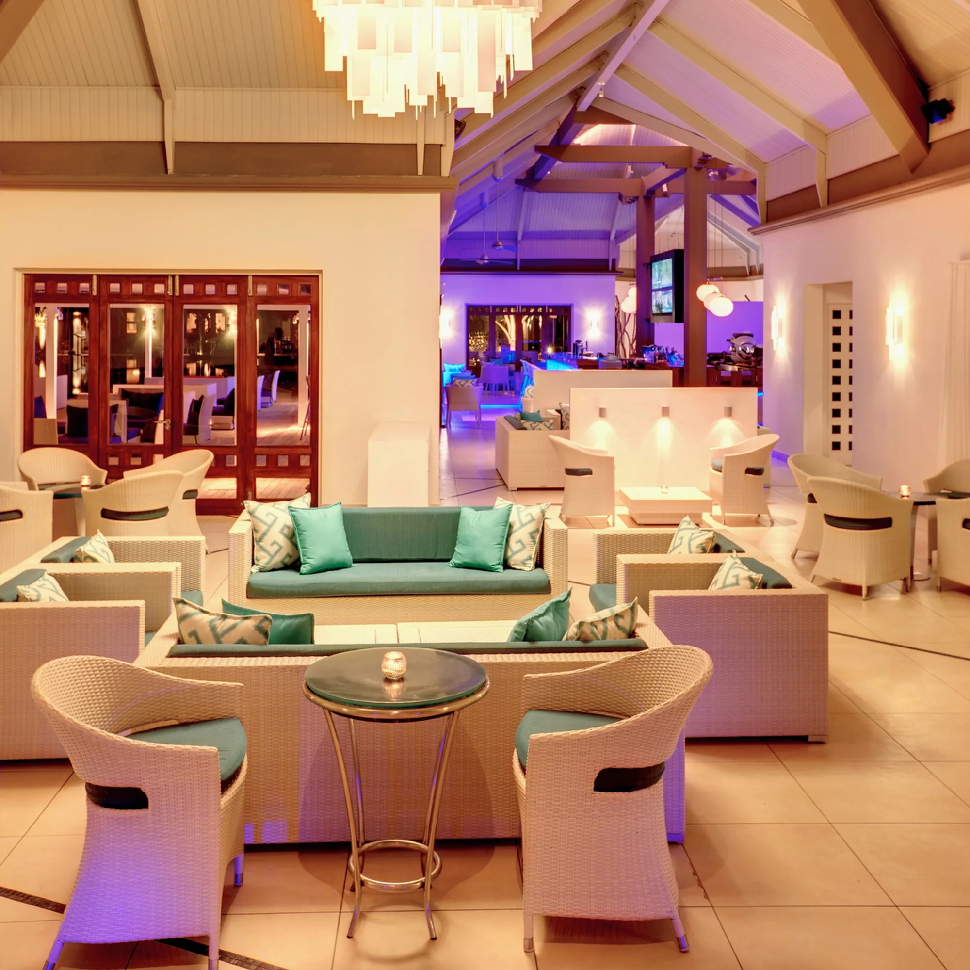 An indoor lounge with sofas, armchairs and a chandelier, lit in purple and gold