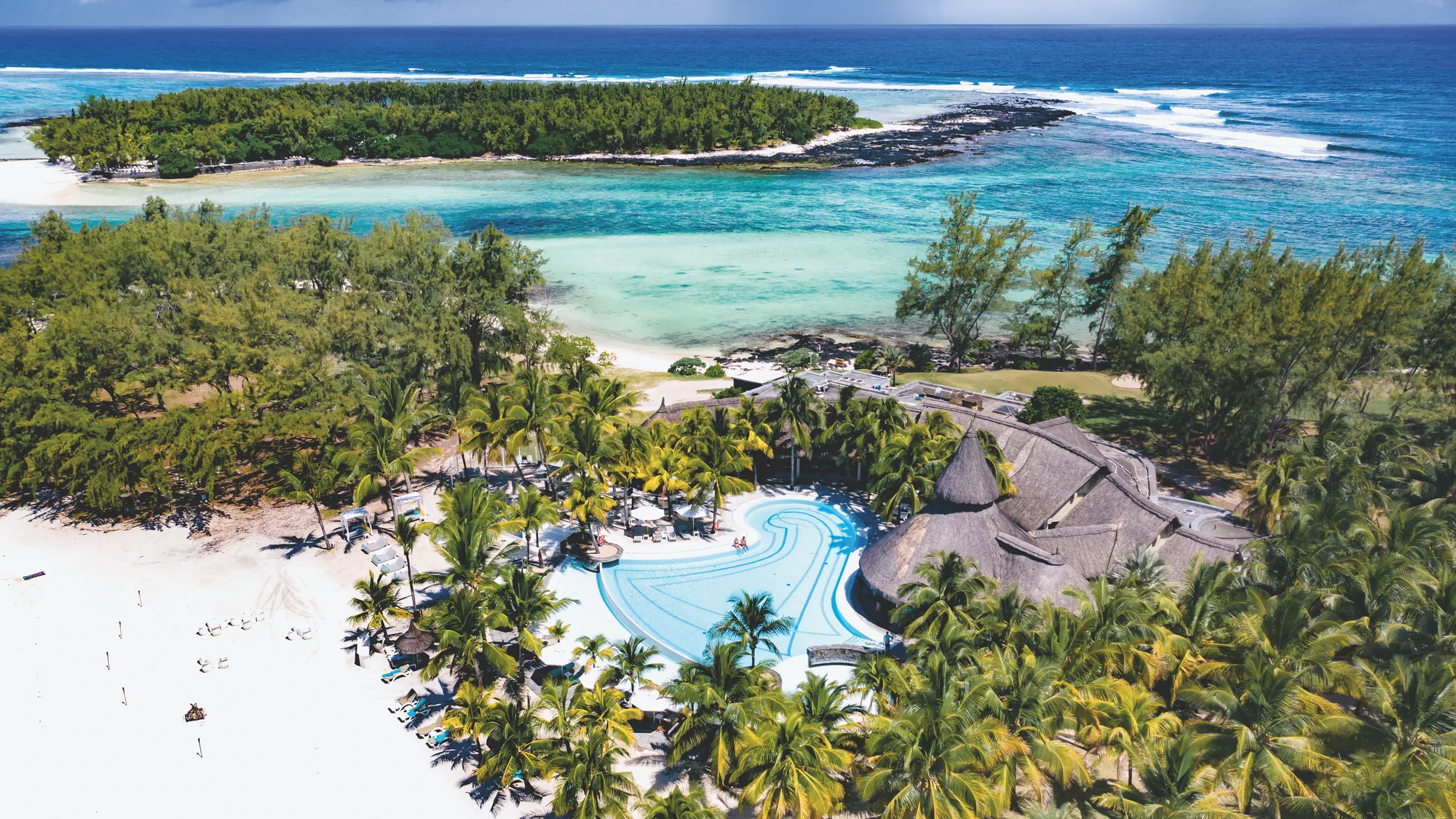 Aerial view of Shandrani Beachcomber Resort & Spa, its pools and thatched roofs beside a turquoise lagoon and a small island