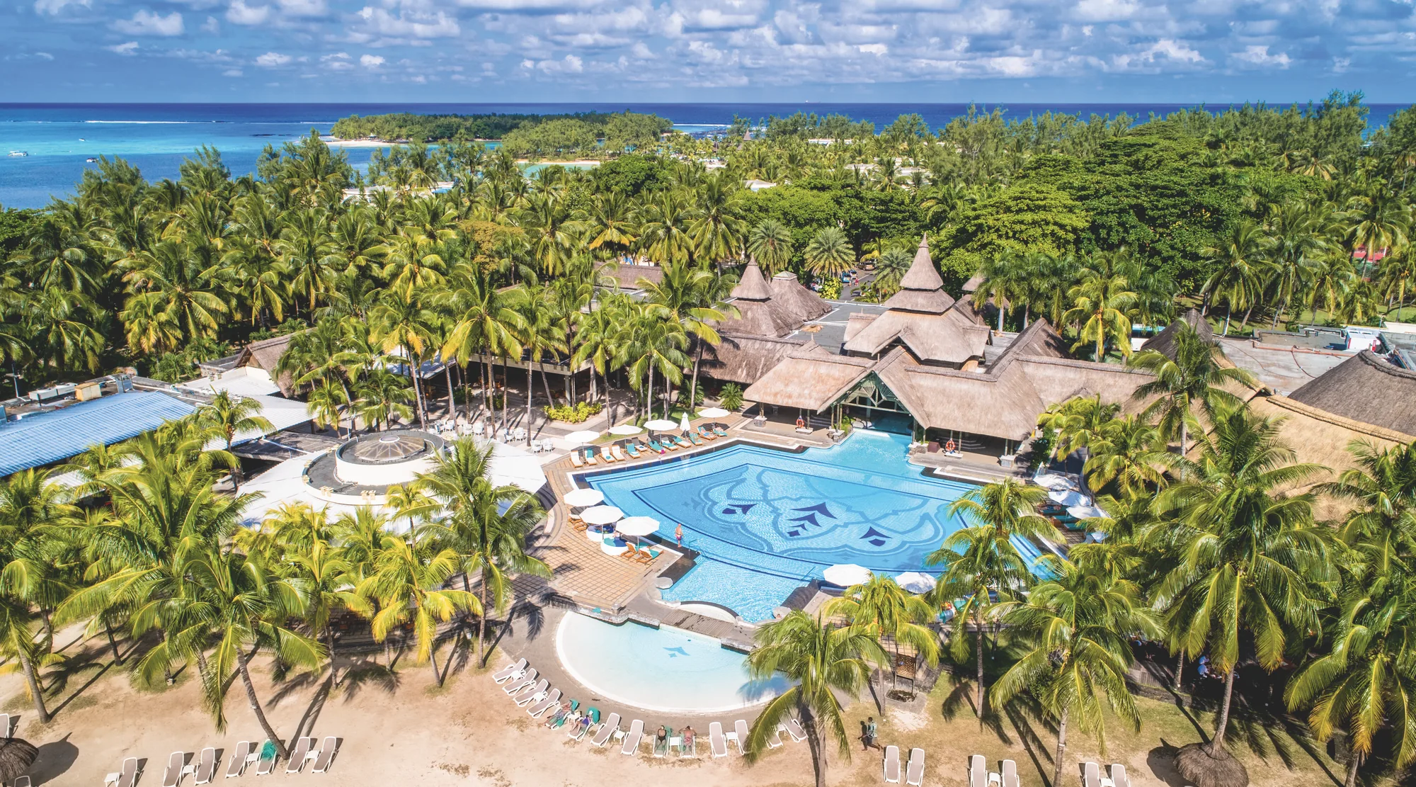 A large pool surrounded by palms and thatched-roof buildings, seen from above