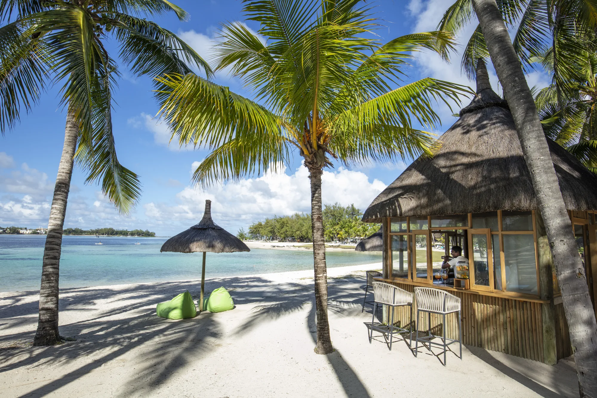 A thatched beach bar with stools on the sand, under palm trees by the lagoon