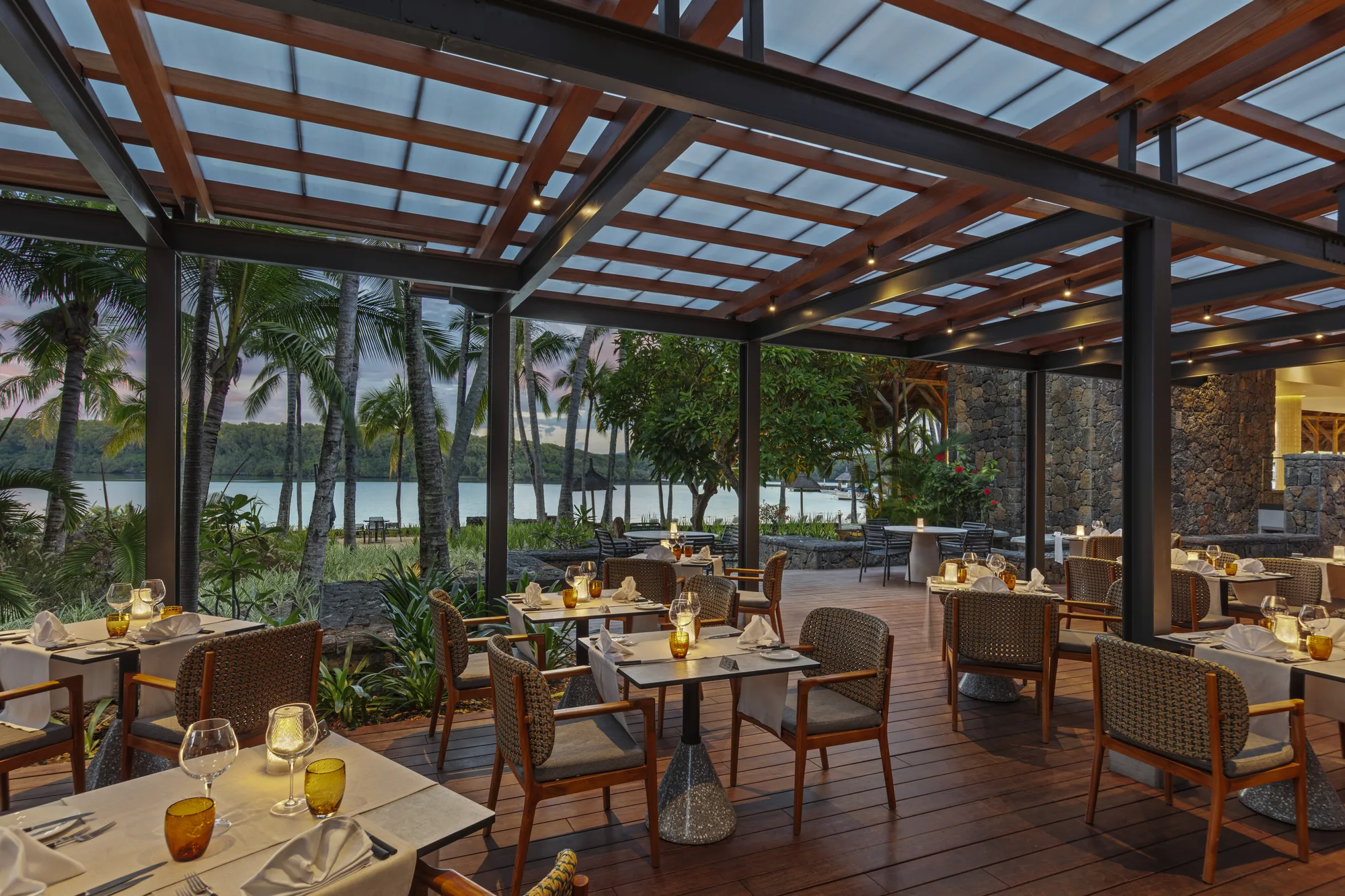 A terrace restaurant under a glass and timber roof, with laid tables and lagoon views