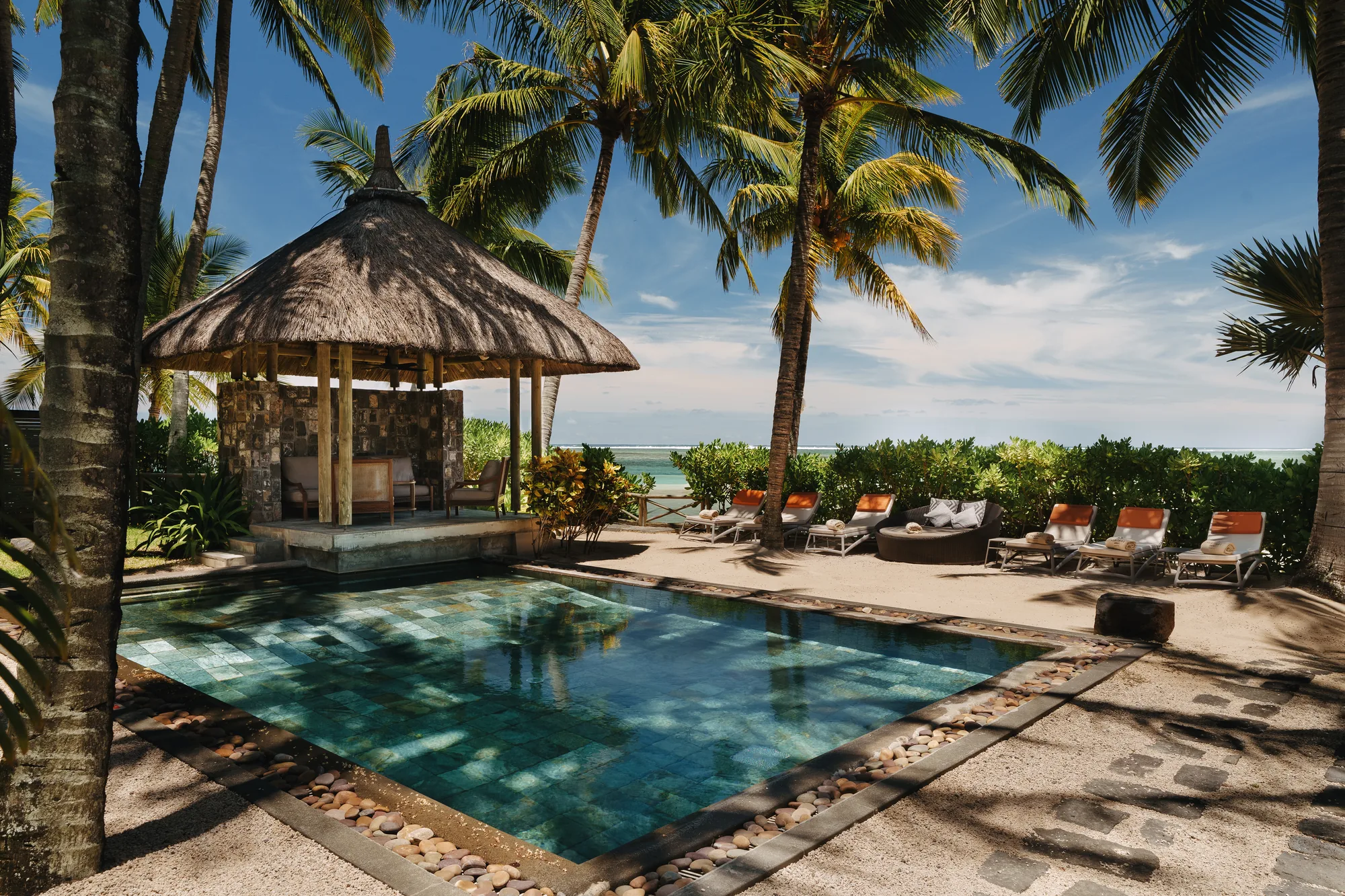 A pool beside a thatched gazebo, with loungers and the sea beyond