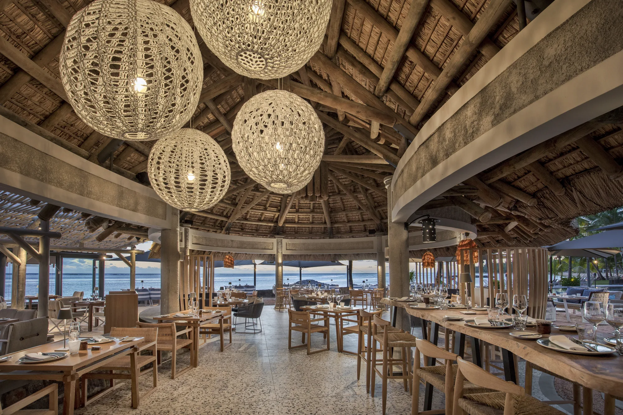 A large thatched restaurant with woven pendant lights and tables open to the sea