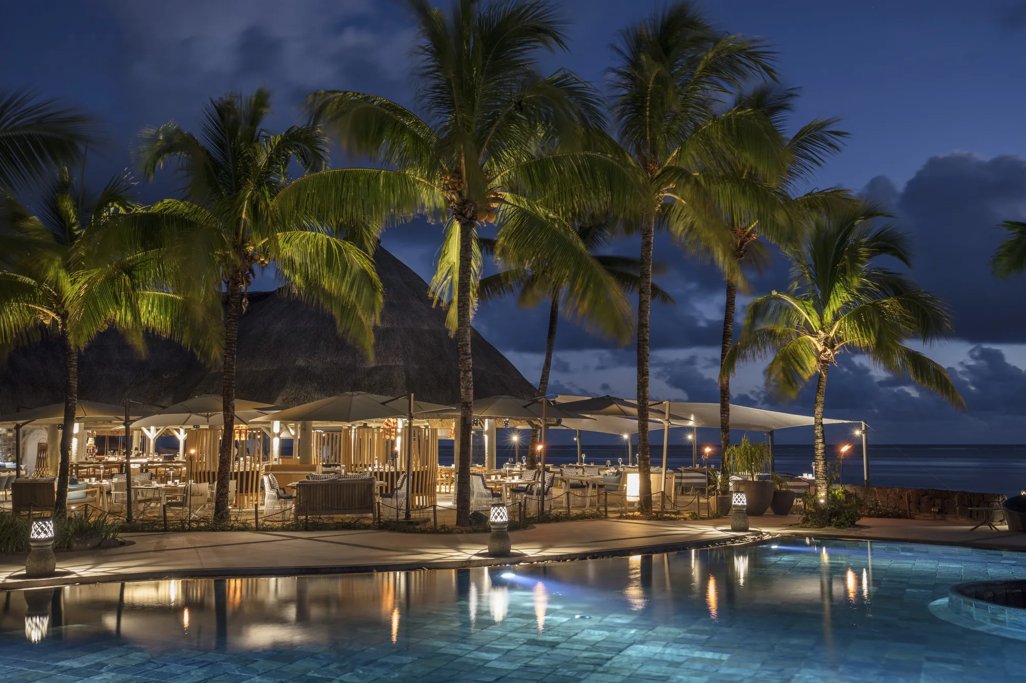 A pool and restaurant under palms, lit at dusk