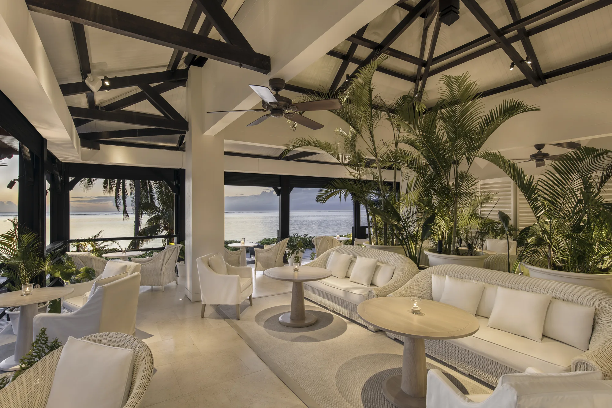 An open lounge with white sofas and potted palms, looking across the lagoon
