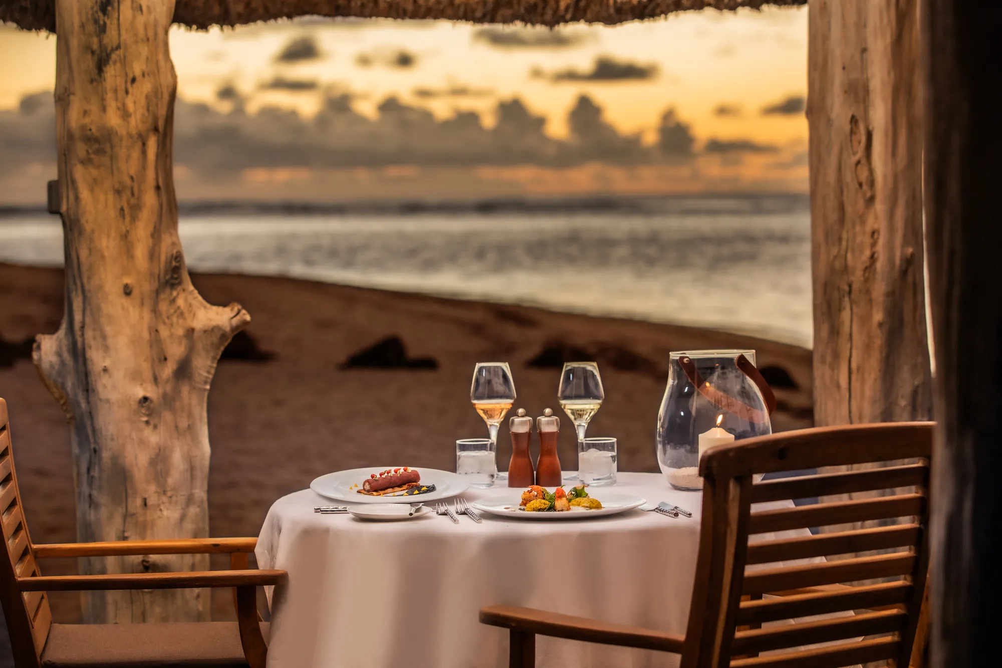 A table laid for two by the sea at sunset, framed by a wooden pavilion