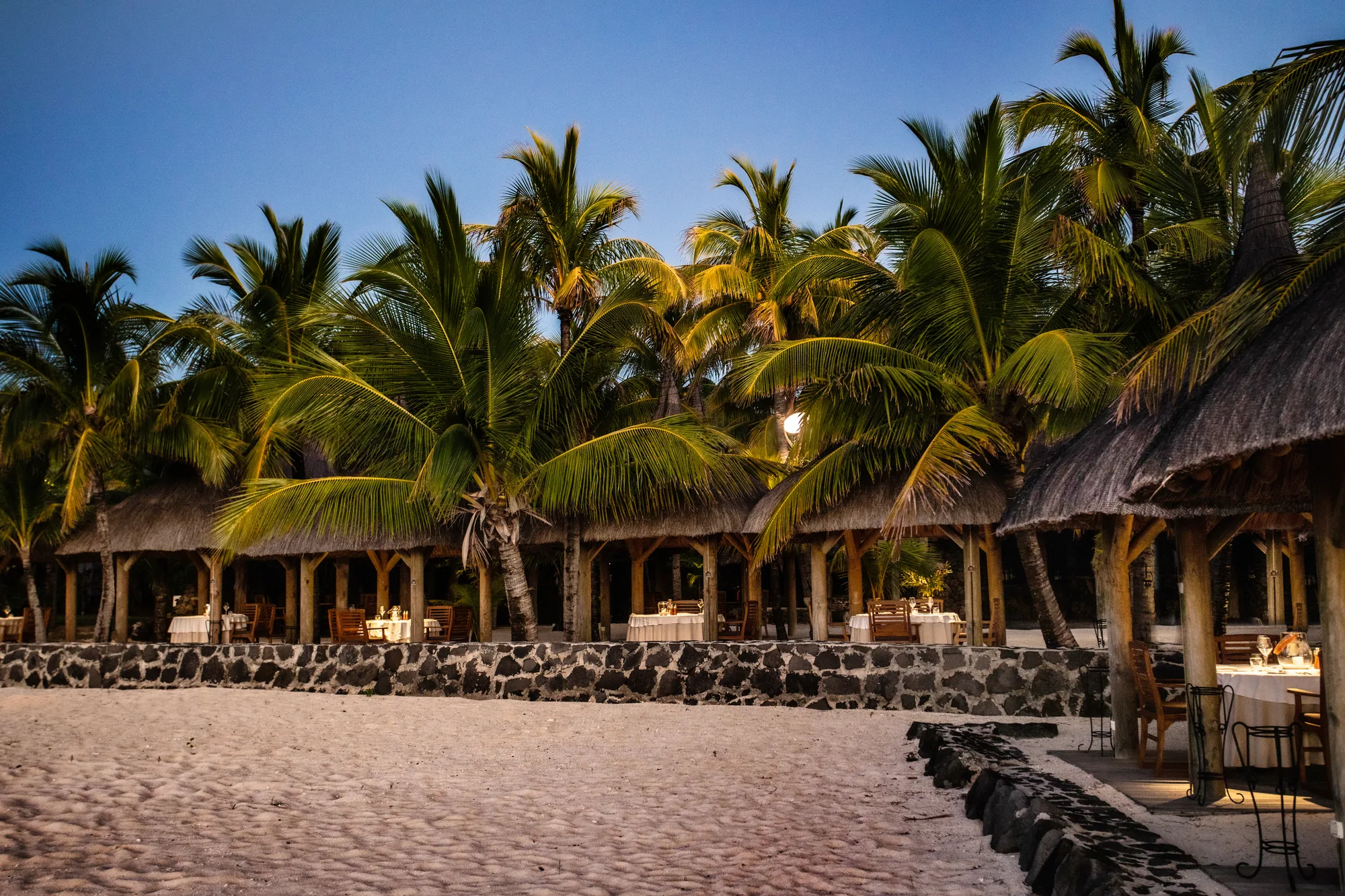 Thatched restaurant pavilions along the beach behind a stone wall, lit at dusk