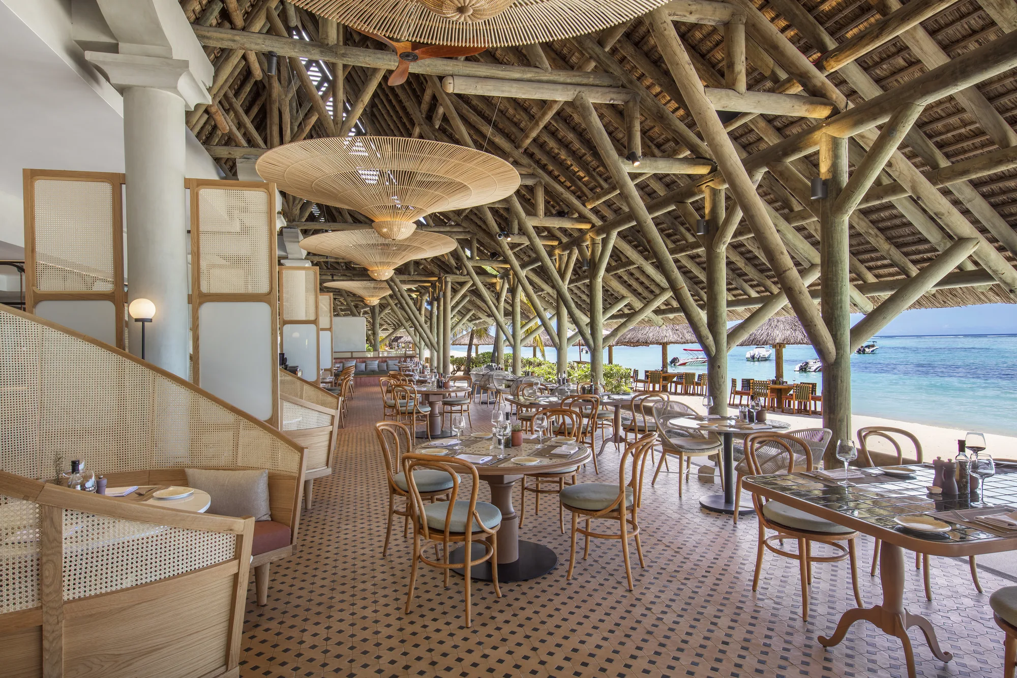 A restaurant under a timber and thatch roof, with rattan pendant lights and tables by the sea