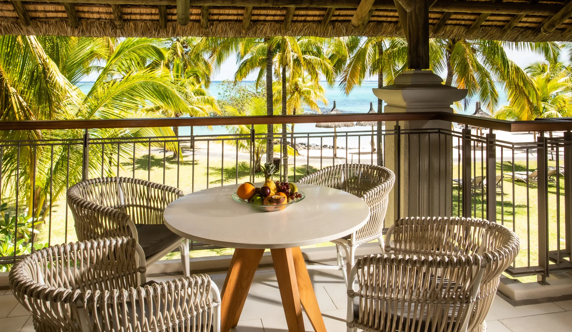 A balcony with woven chairs and a round table, looking over palms to the beach