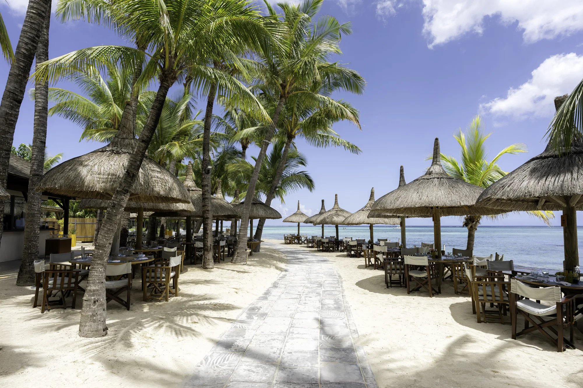 Restaurant tables under thatched parasols along a sandy path by the sea
