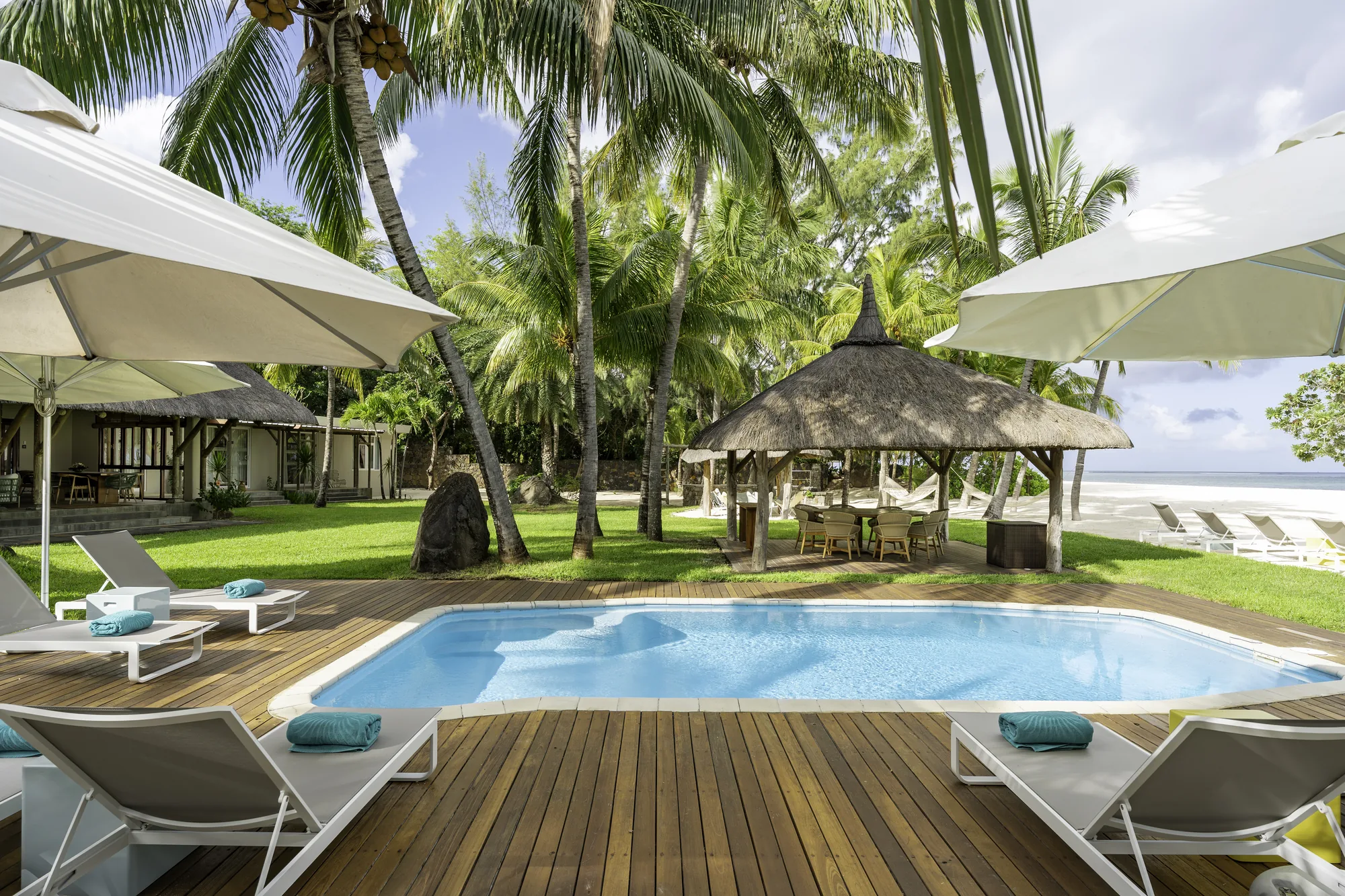 A pool on a wooden deck beside a thatched gazebo, with loungers on the lawn