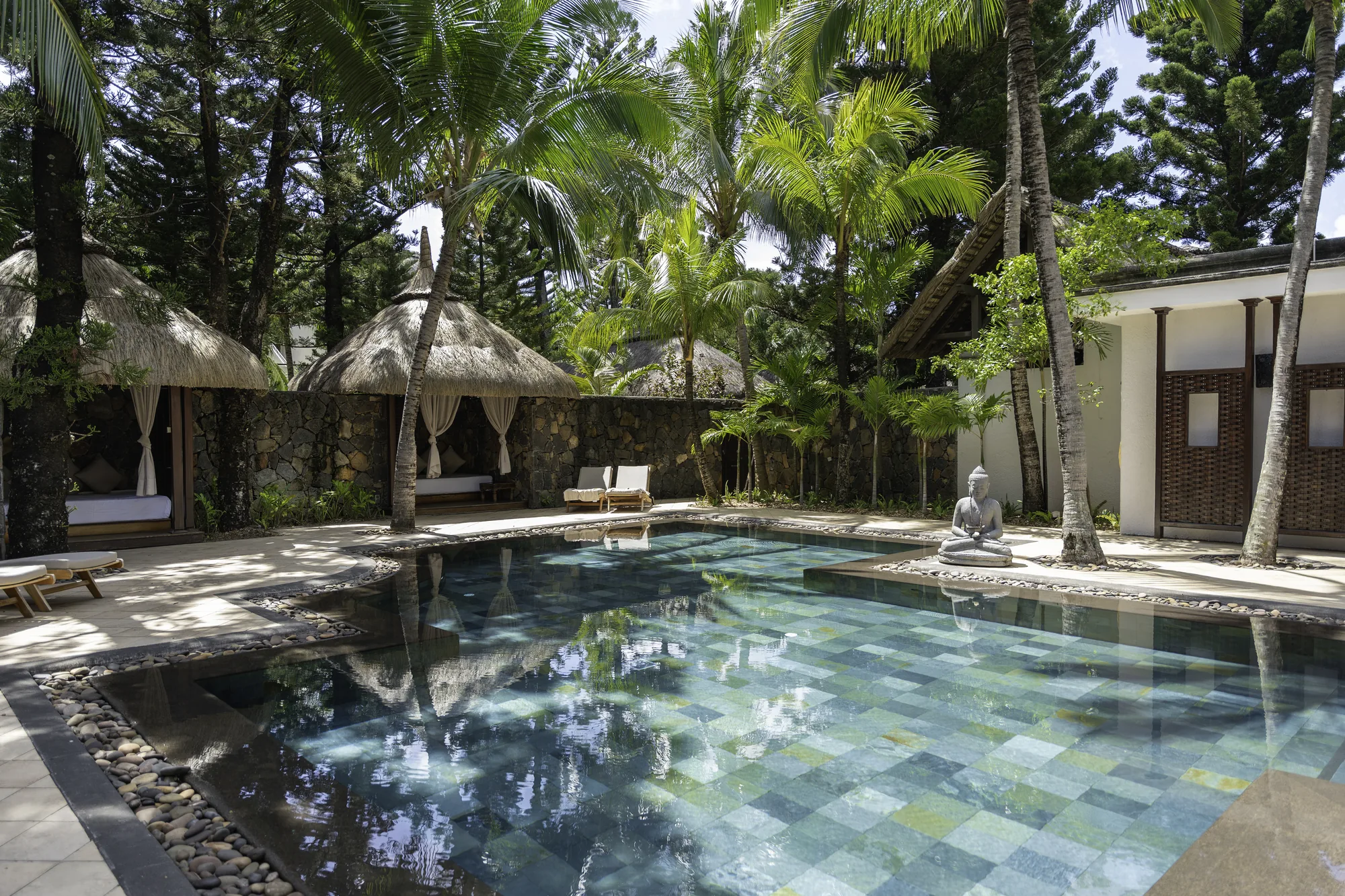 A green-tiled pool with a seated Buddha statue at its edge, beside curtained thatched pavilions under palm trees