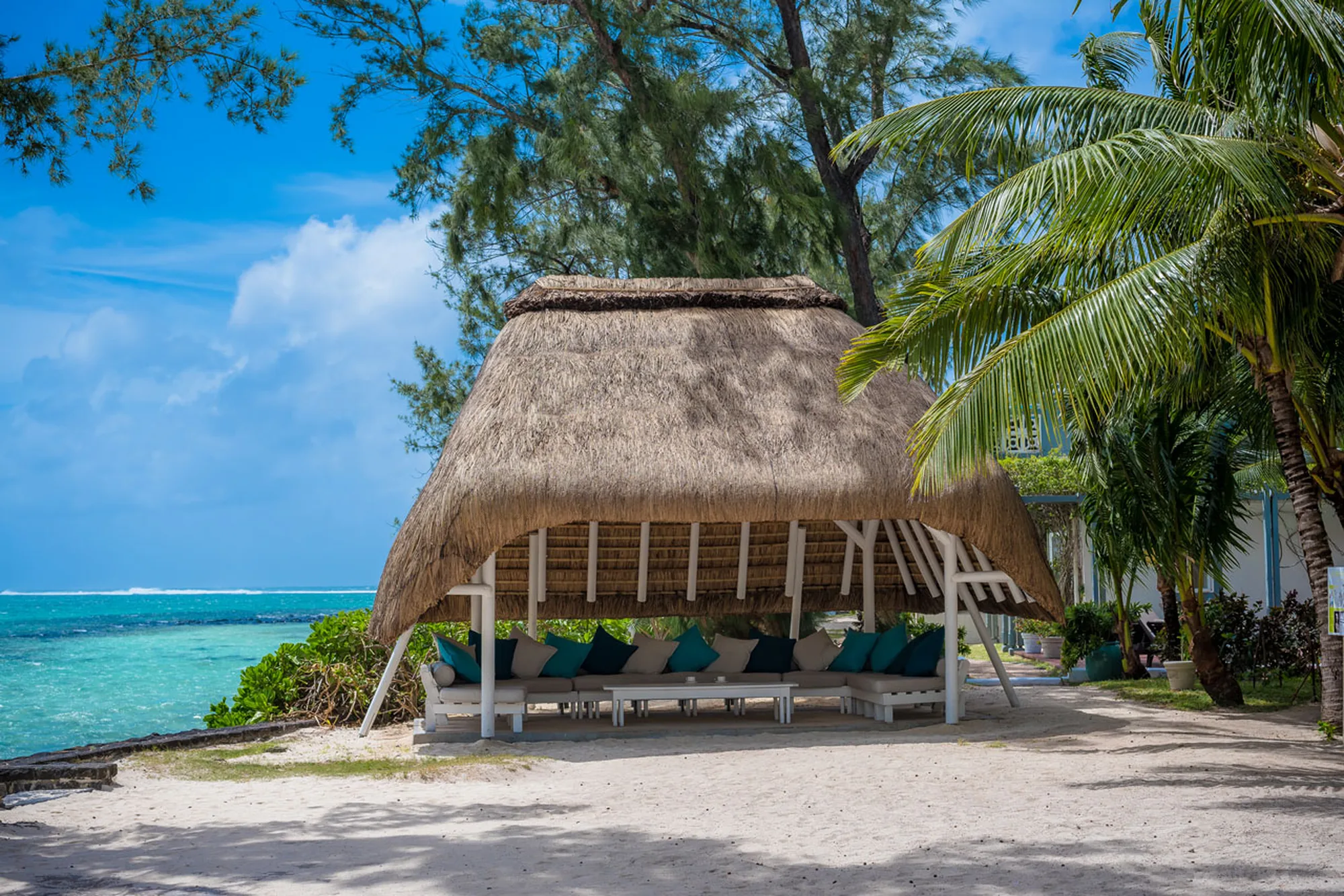 A thatched pavilion with cushioned seating on the sand beside the sea