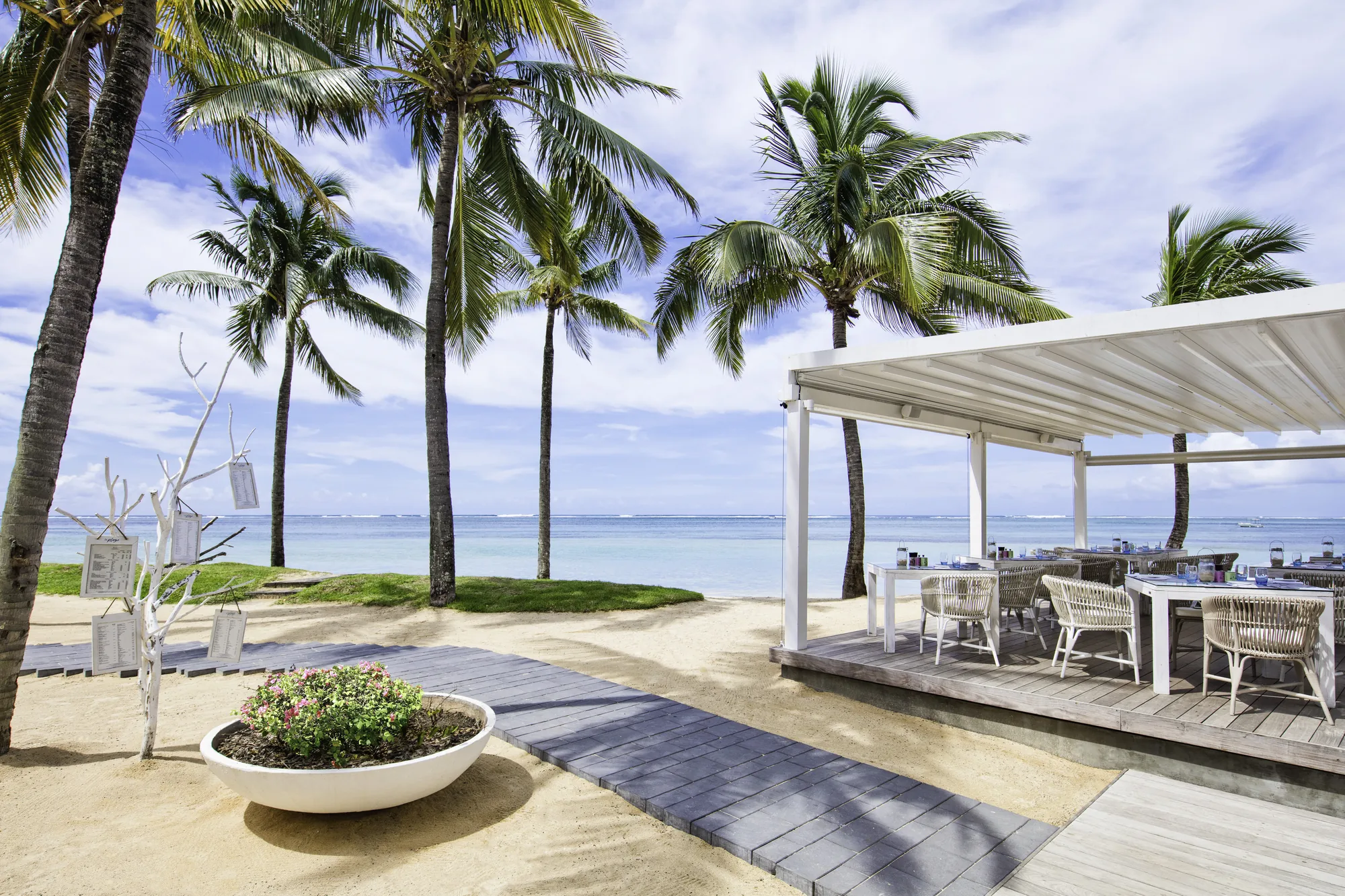A covered beach deck with tables and chairs, under palm trees by the sea