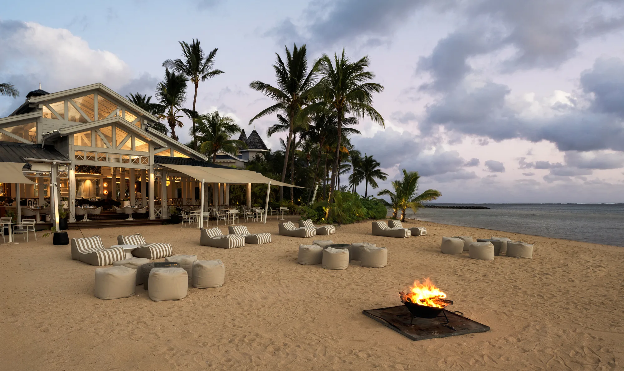 The beach at dusk, with a fire pit, low seating and a restaurant behind