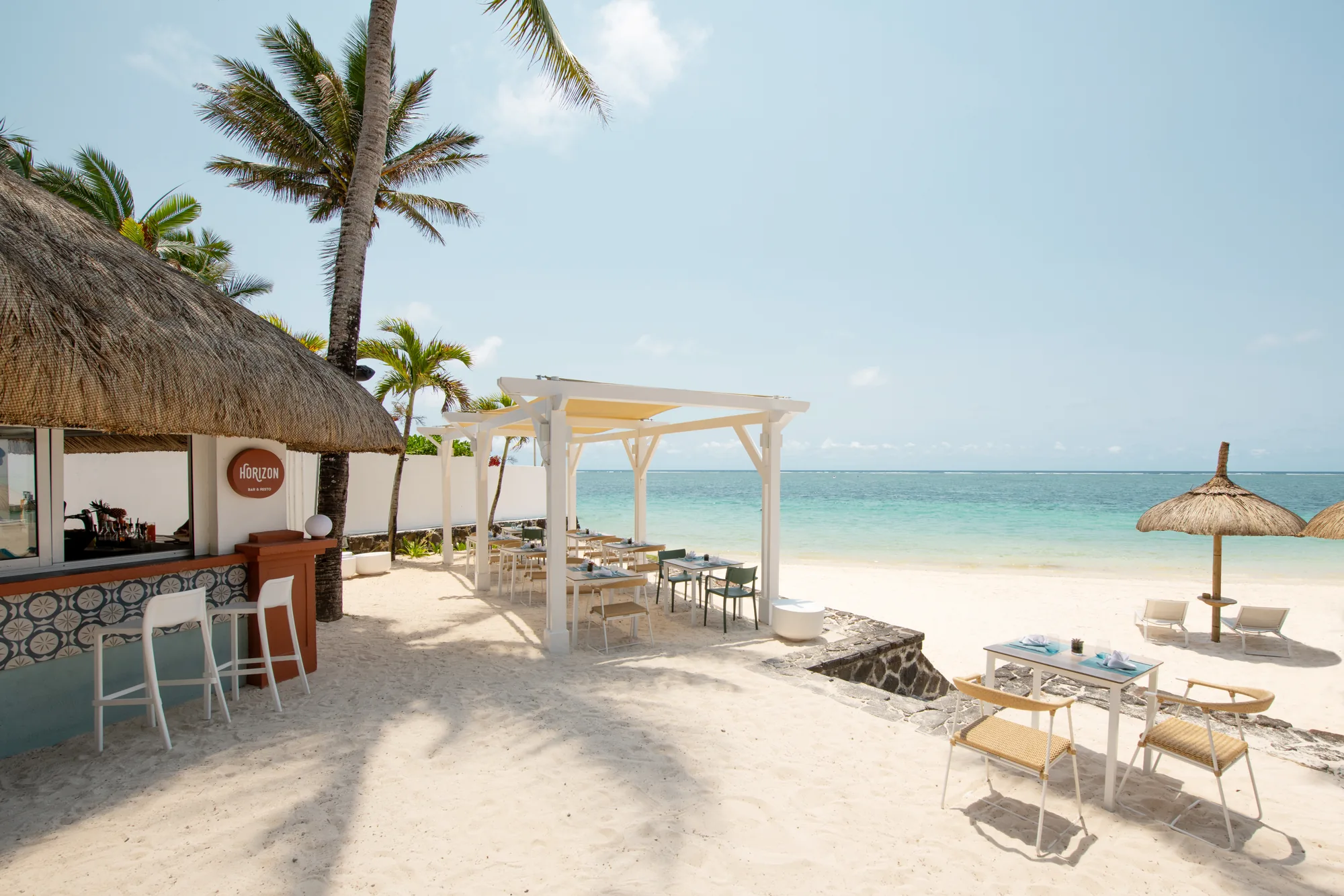 A beach bar and tables on the sand, with a pergola and the sea beyond