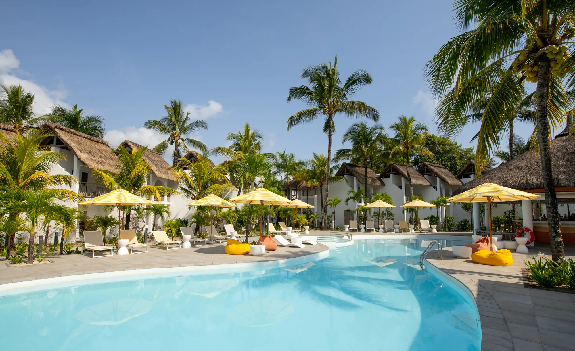 A curved pool with yellow parasols and beanbags, in front of thatched accommodation