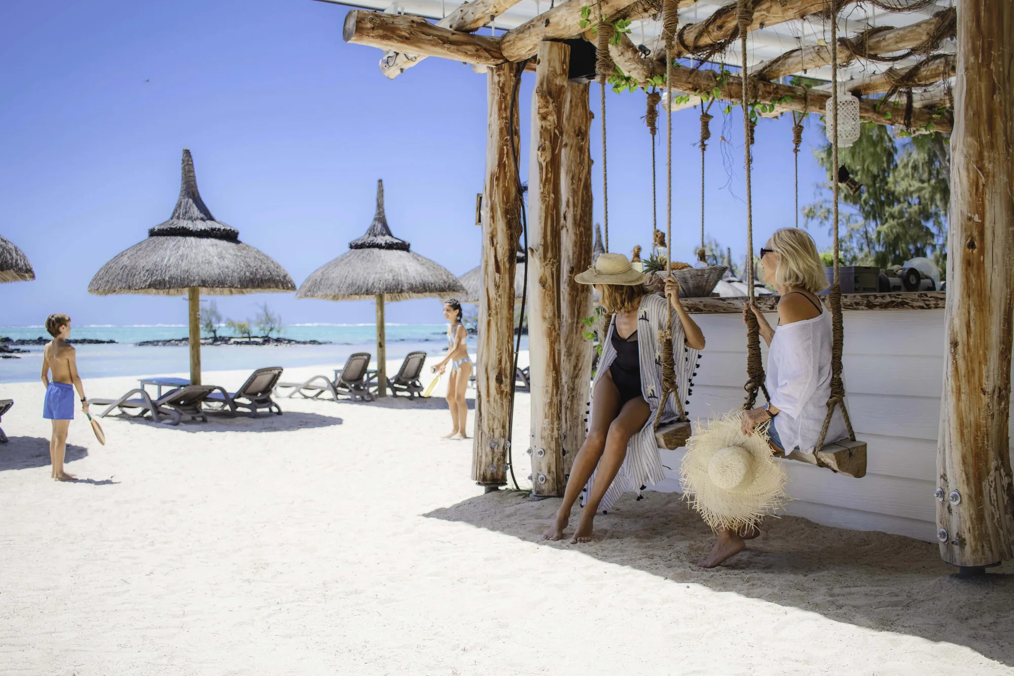 Two guests on swing seats at a beach bar, with thatched parasols on the sand