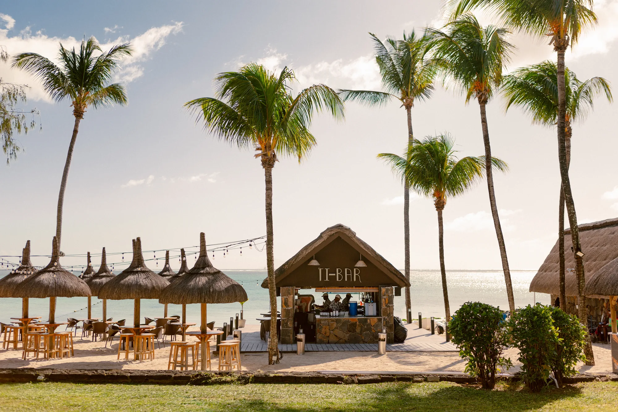 A thatched beach bar and parasol-shaded tables on the sand, among palm trees