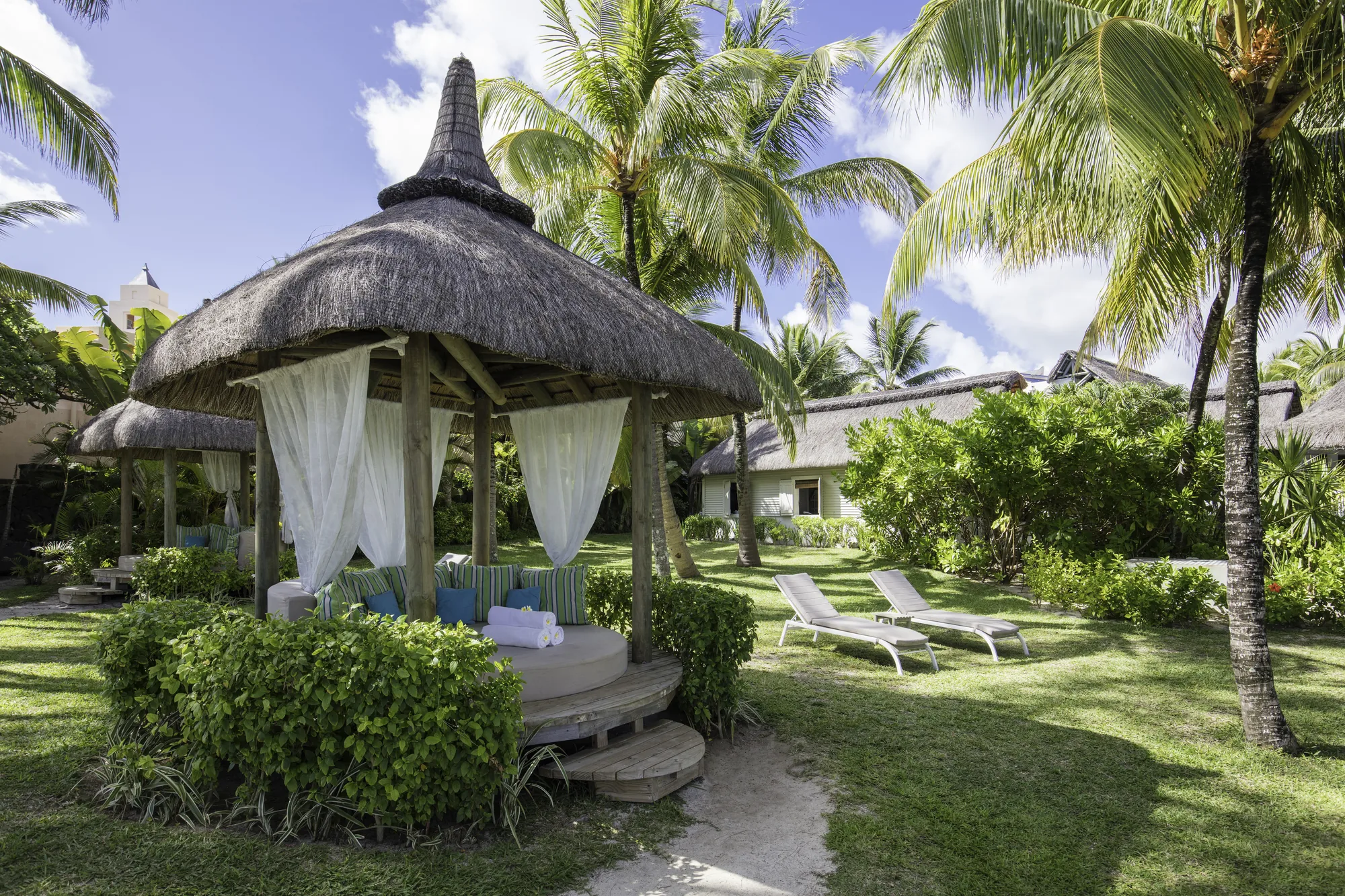 A thatched gazebo with white curtains on a lawn among palm trees