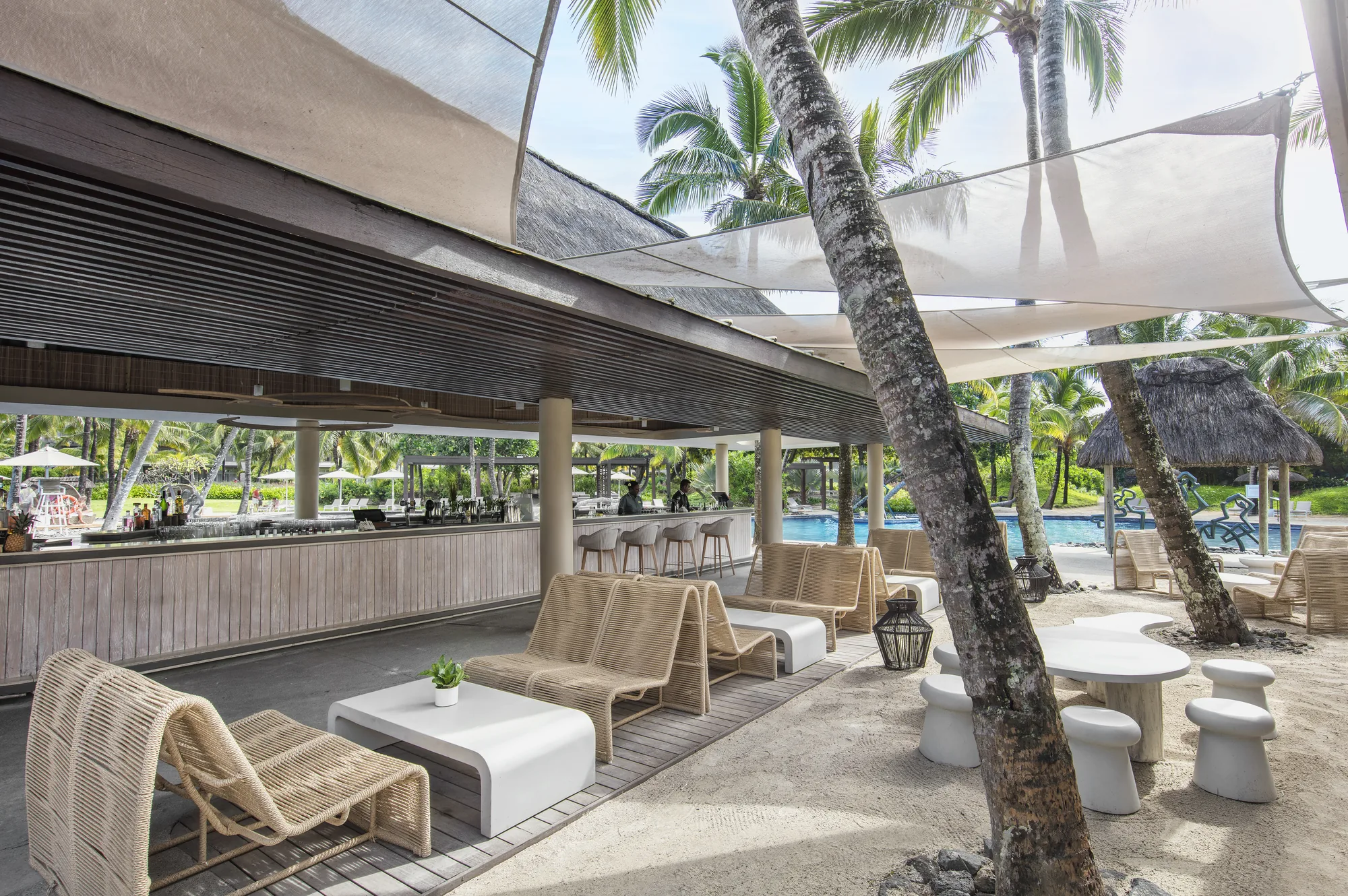 A shaded beach bar with woven loungers and low tables under palm trees