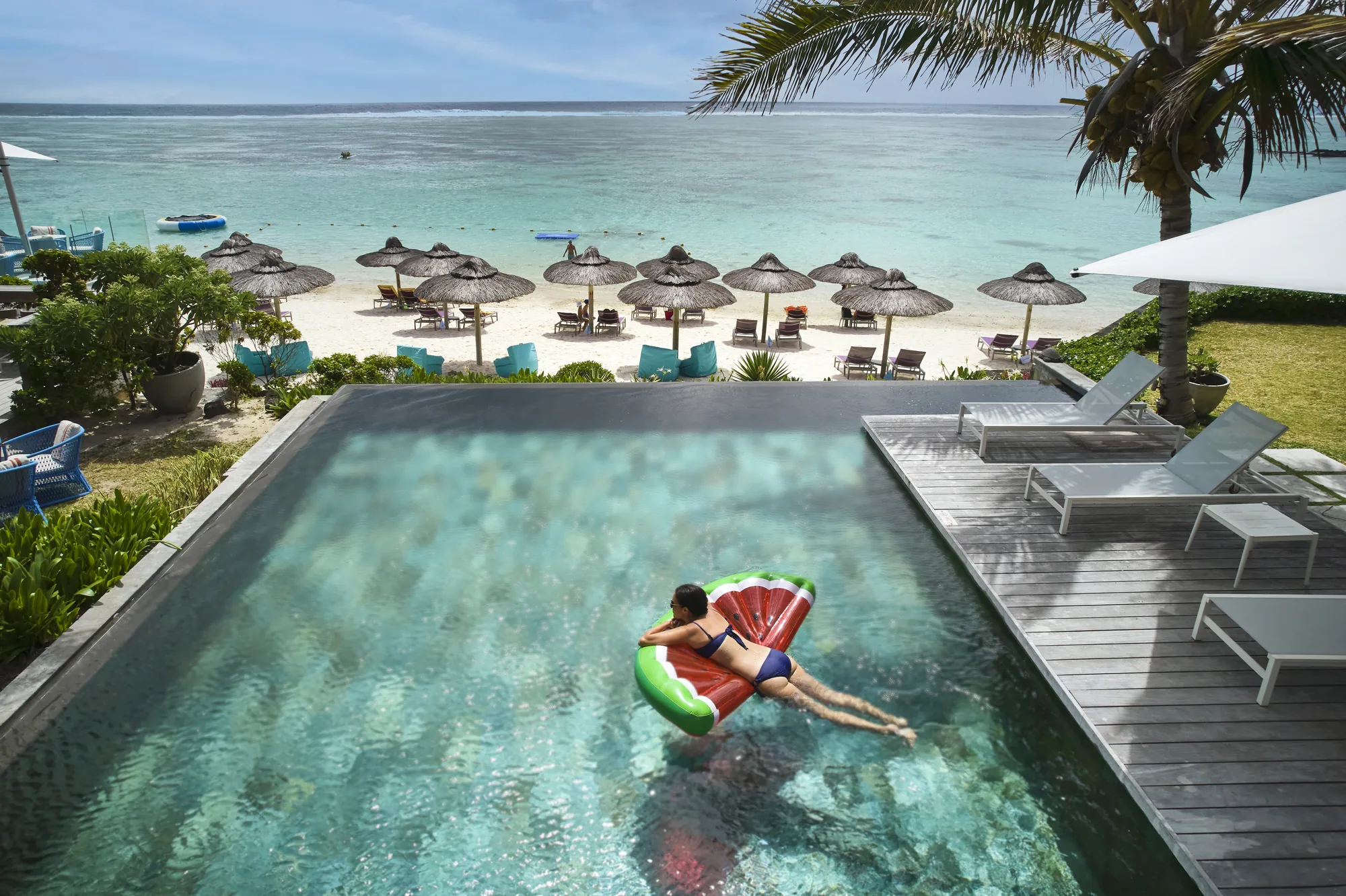 A guest on a watermelon float in a pool, with the beach and parasols beyond