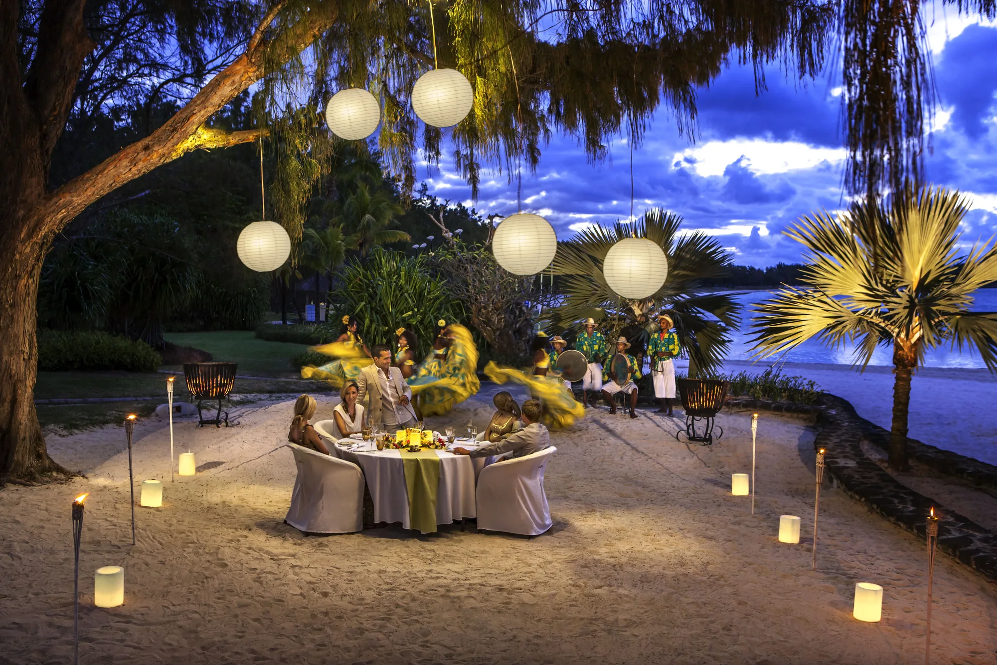 A candlelit dinner on the beach at night, with paper lanterns hung in the trees and musicians nearby