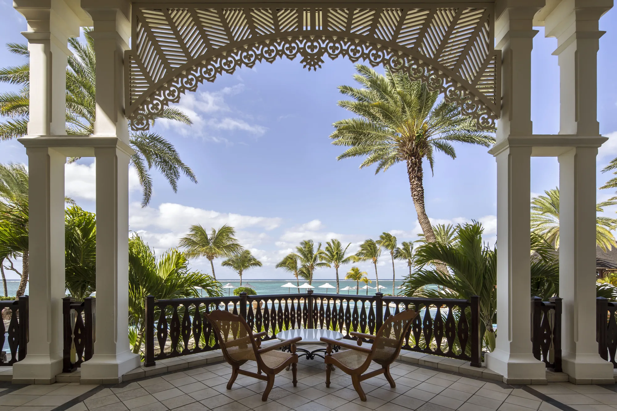 A balcony with carved fretwork and two chairs, overlooking palm trees and the sea