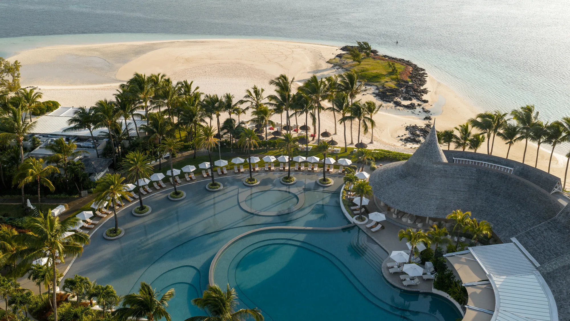 Aerial view of a large pool, a thatched building and the beach and lagoon