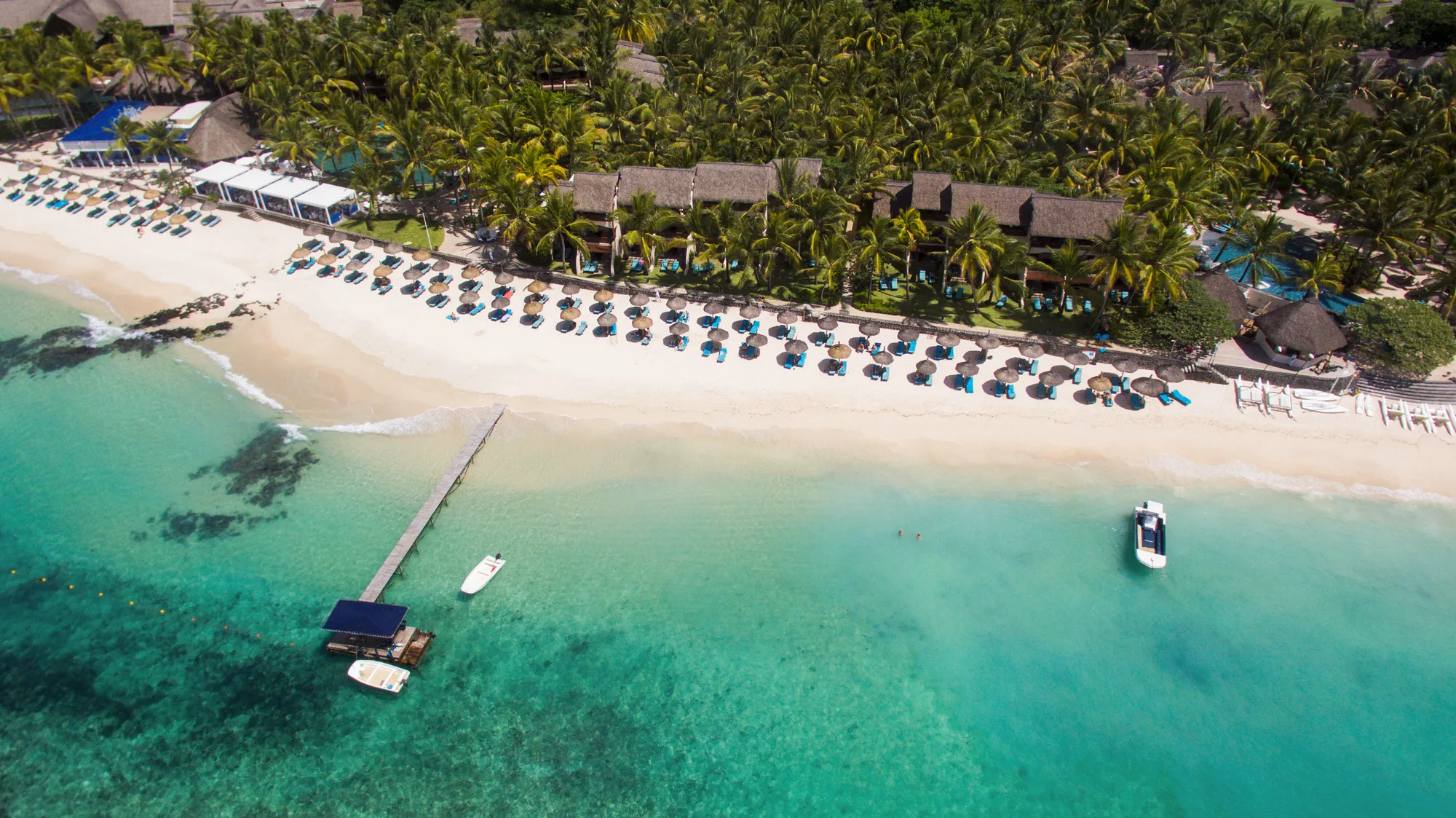 Aerial view of Constance Belle Mare Plage, its long beach, jetty and parasols beside a turquoise lagoon