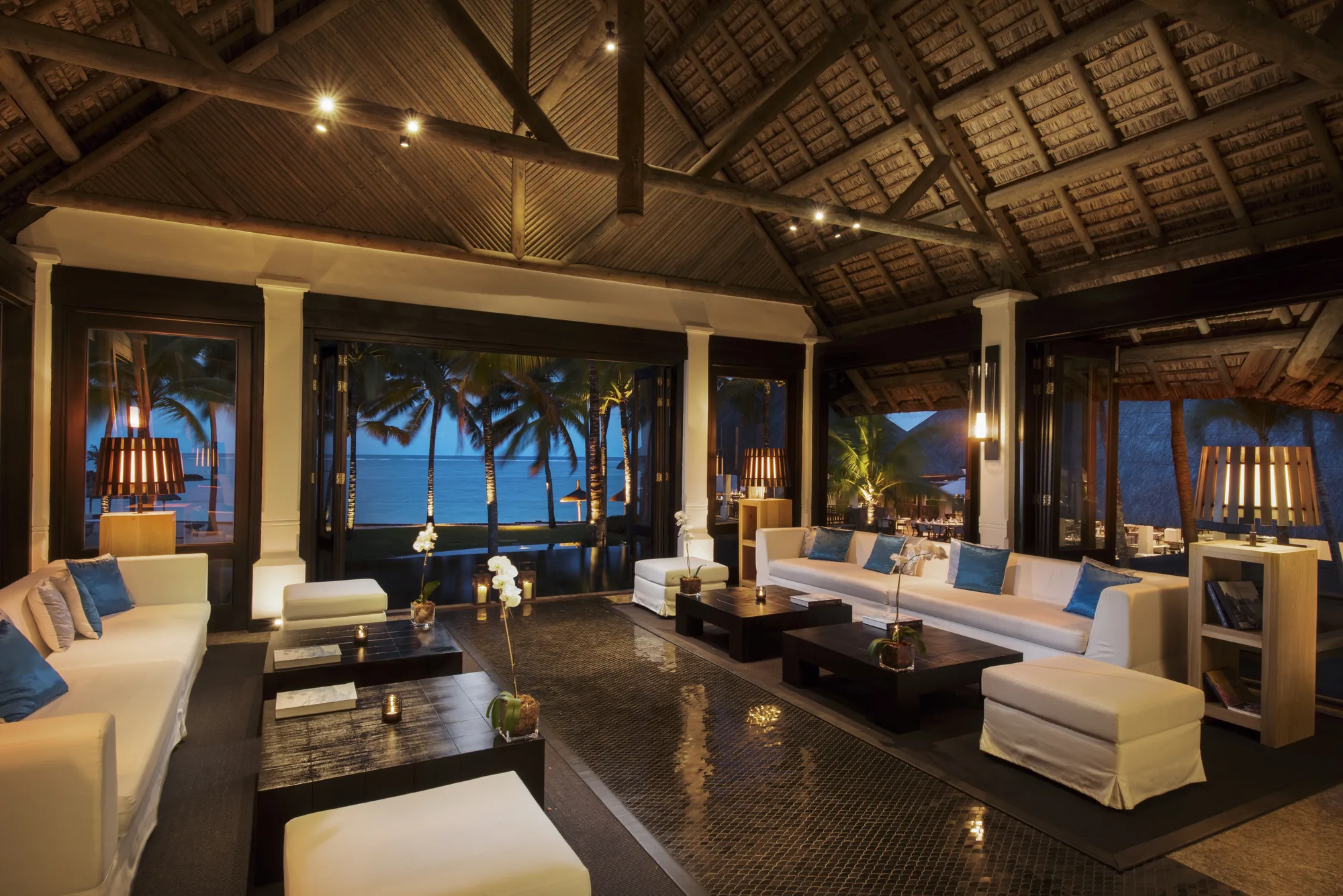 A lounge with white sofas under a timber roof, open to the sea at night