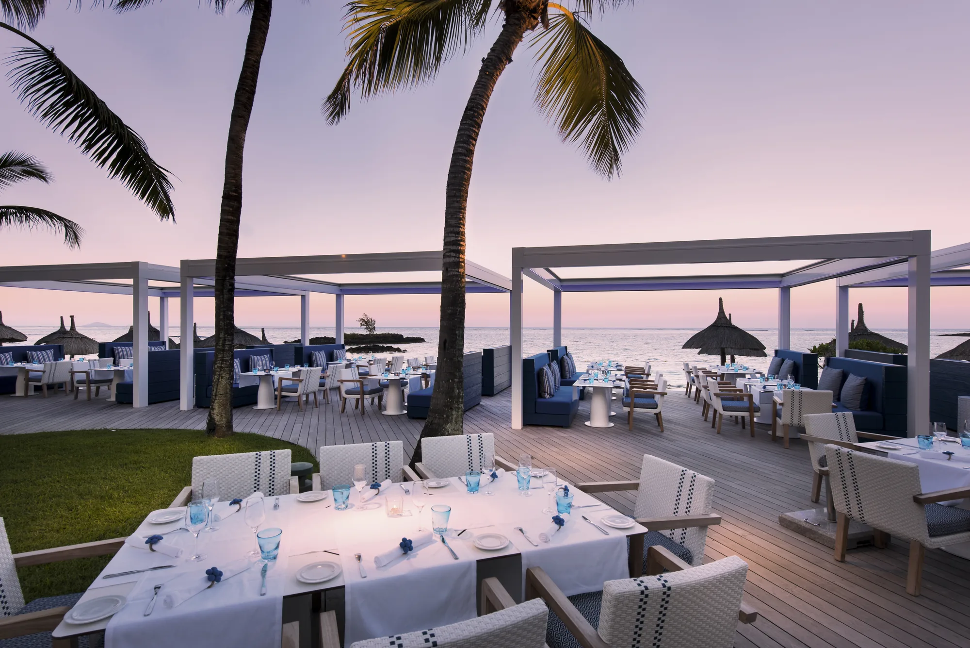 A seafront deck restaurant with tables laid under pergolas at dusk
