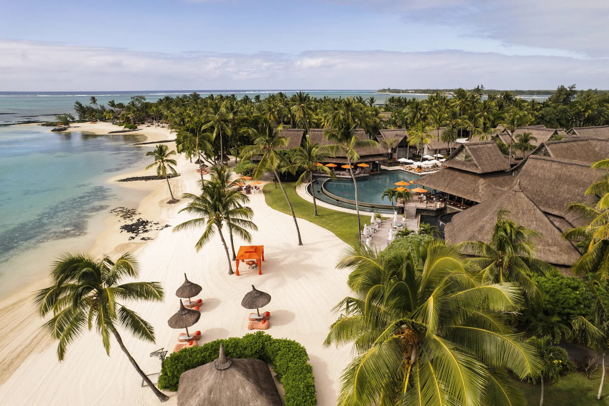 A white beach with thatched parasols beside a pool and thatched-roof buildings