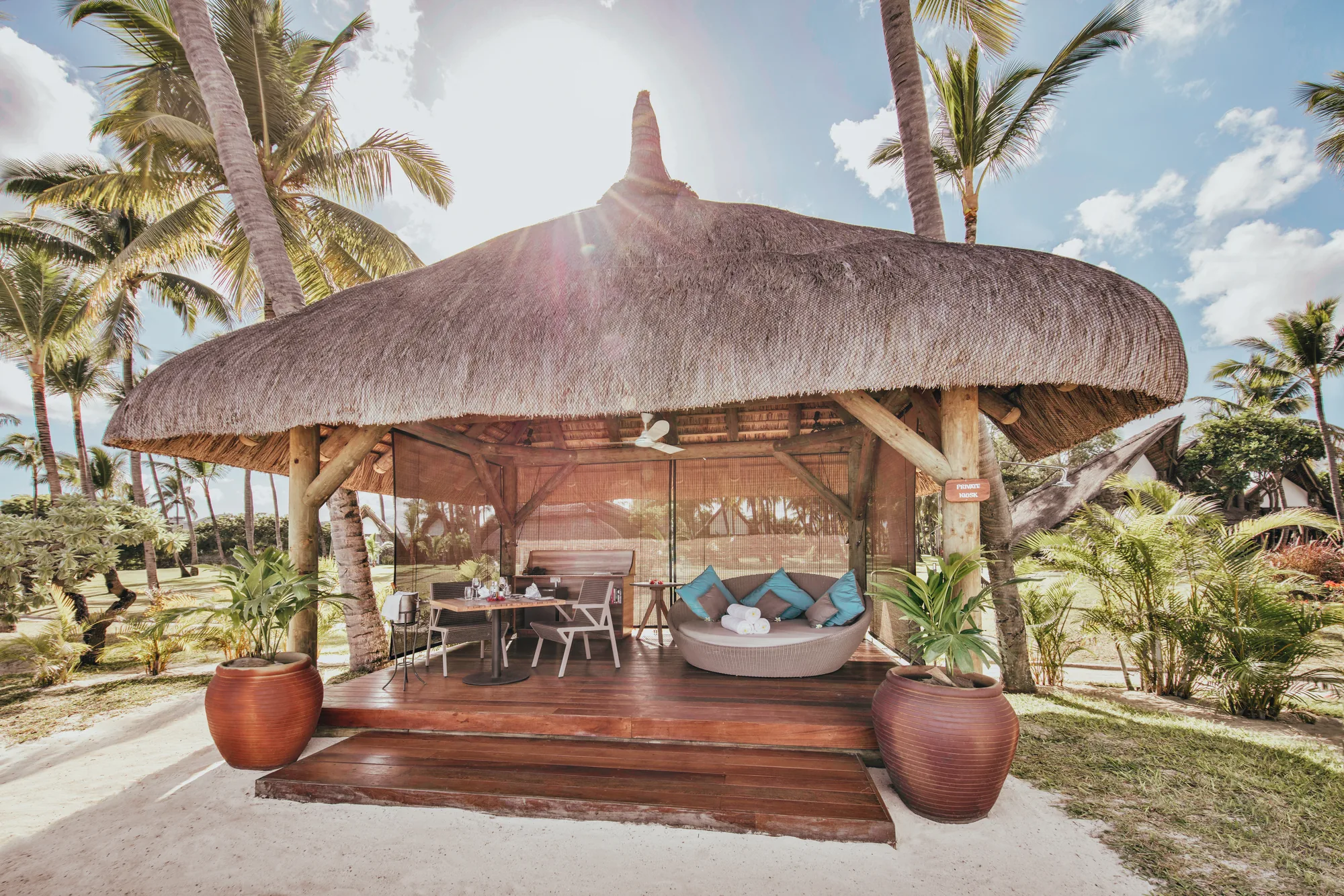 A thatched gazebo with a daybed and a dining table on a wooden deck