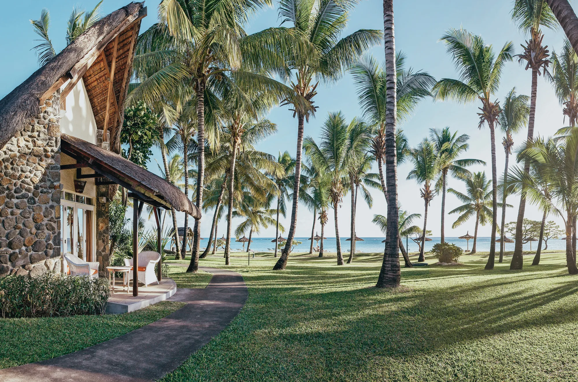 A thatched cottage with a stone wall, and a path through palm trees to the sea