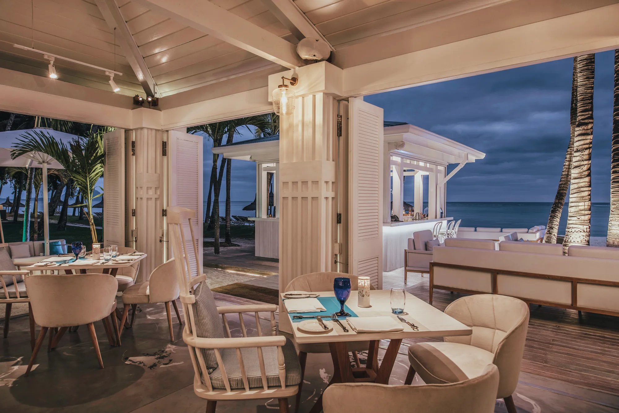 A restaurant with laid tables under a white timber roof, open to the sea