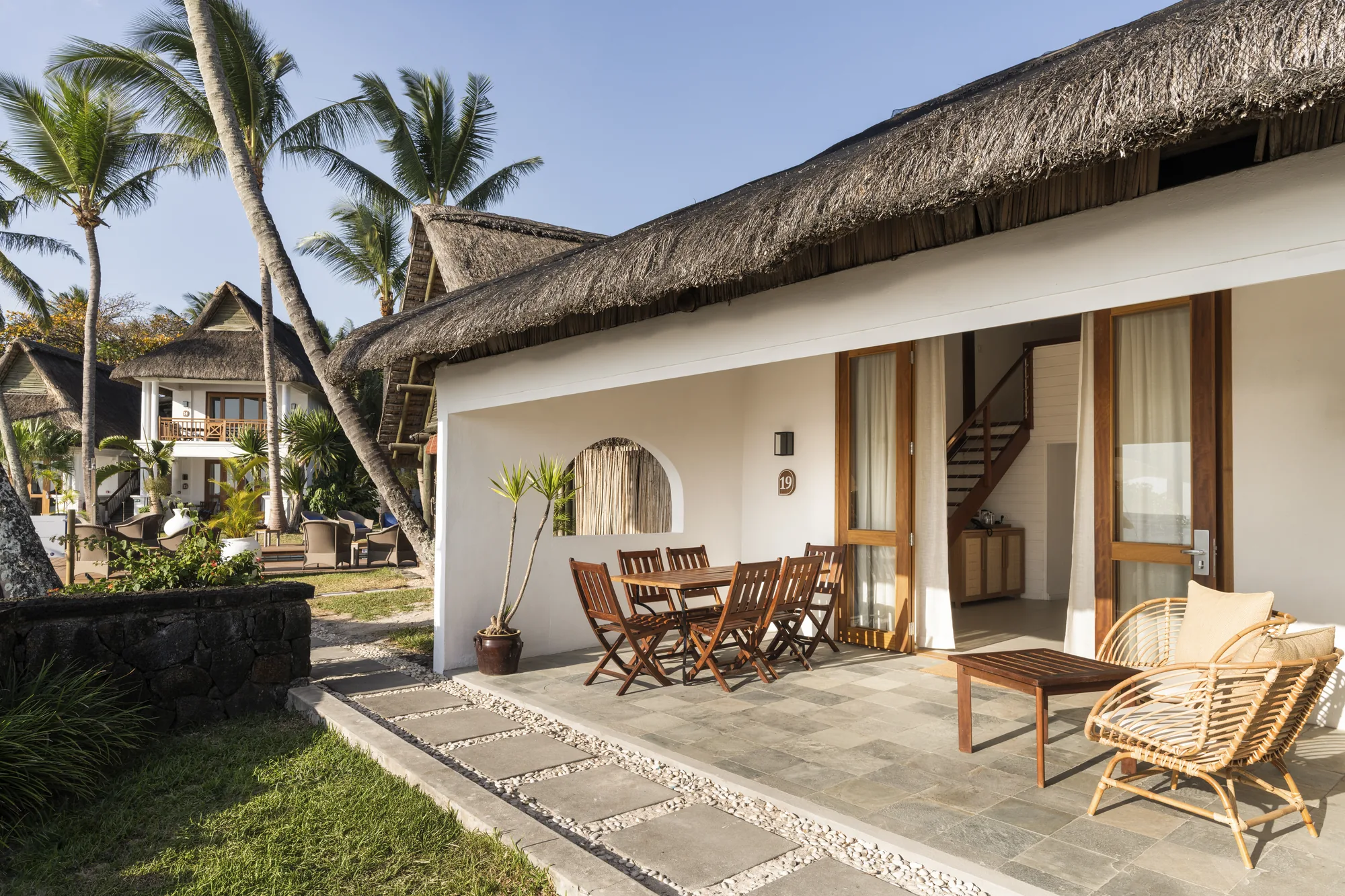 A terrace with a dining table and rattan armchairs beneath a thatched roof
