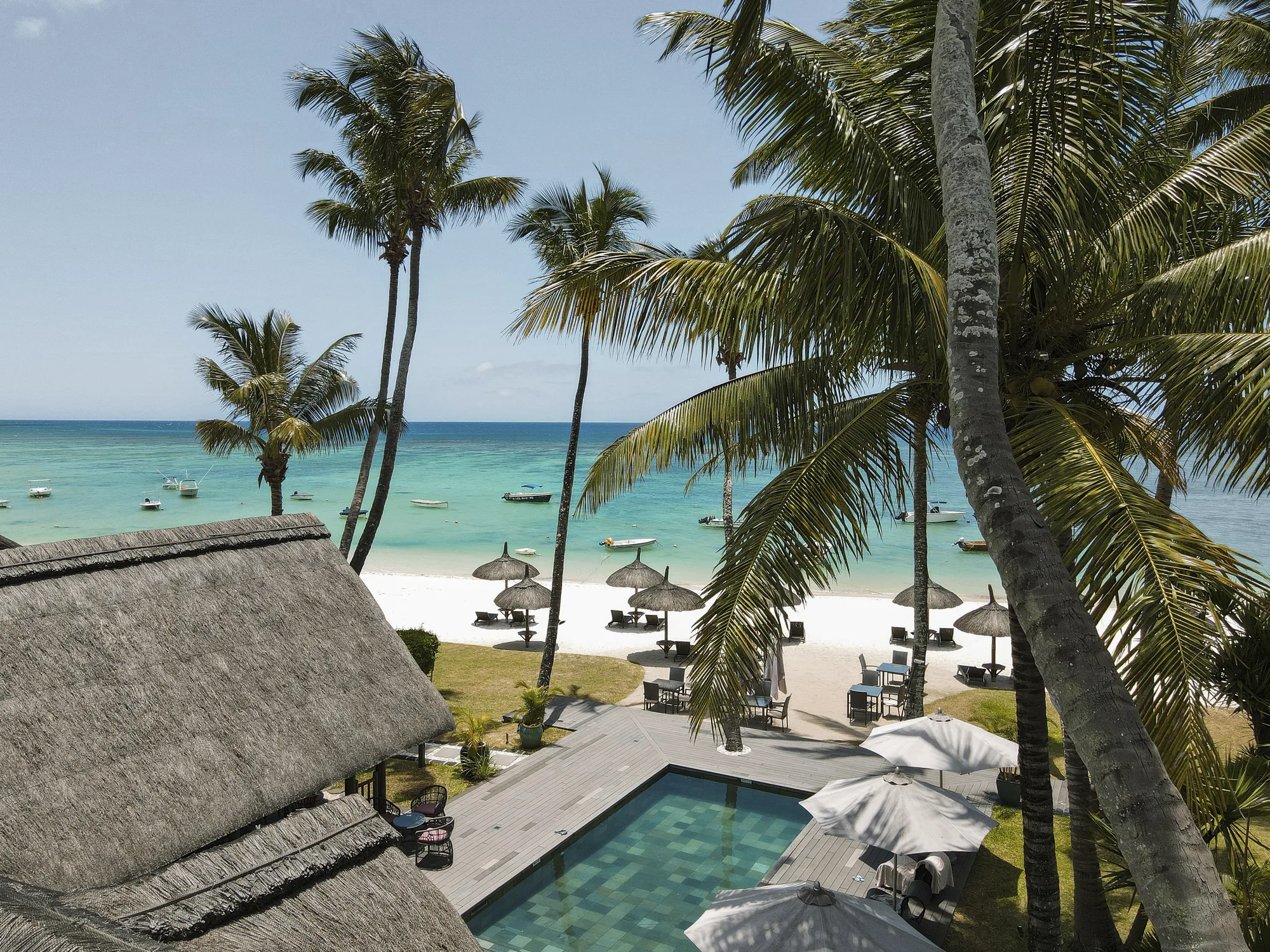 Palm trees, a pool and thatched parasols on the beach, seen from a balcony above