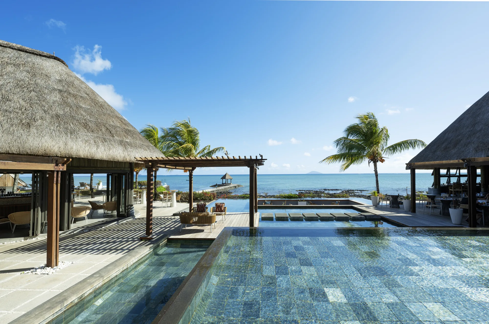 A tiled pool between thatched pavilions, with a pergola and the sea beyond