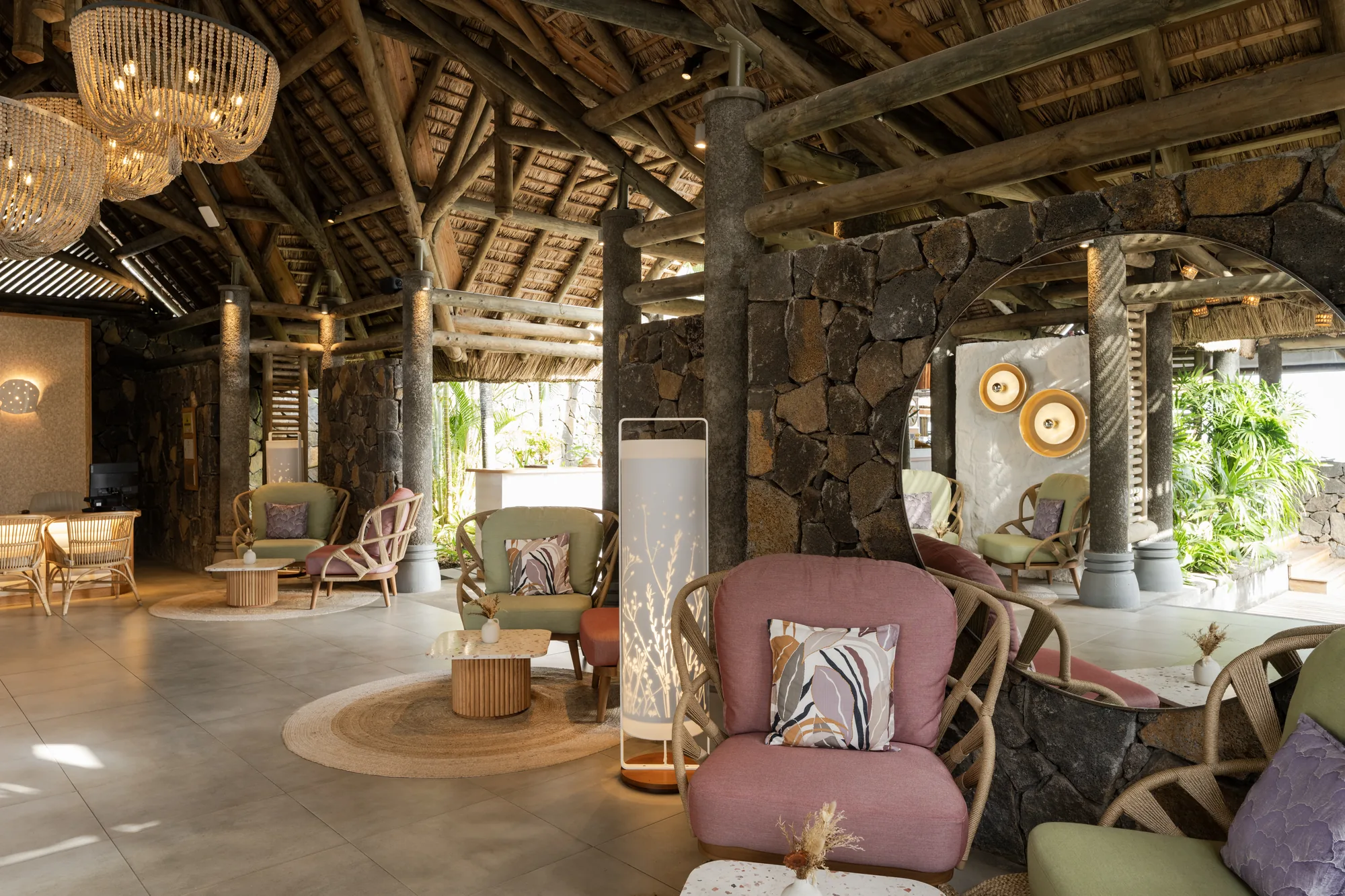 A lounge with pink armchairs, stone walls and woven chandeliers under a thatched roof